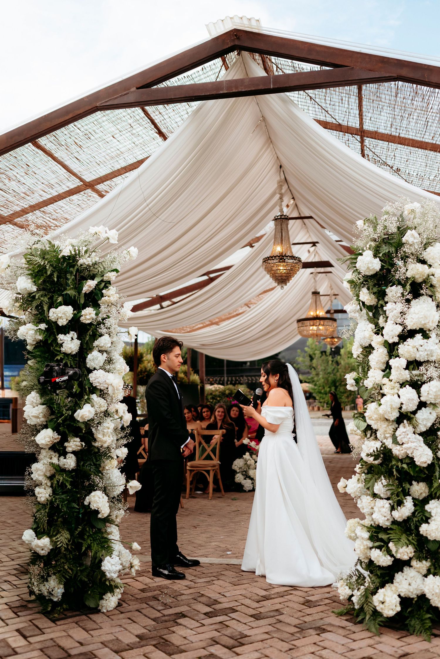 Bride and groom exchange vows under a floral arch with white roses and draped fabric at an outdoor wedding ceremony.