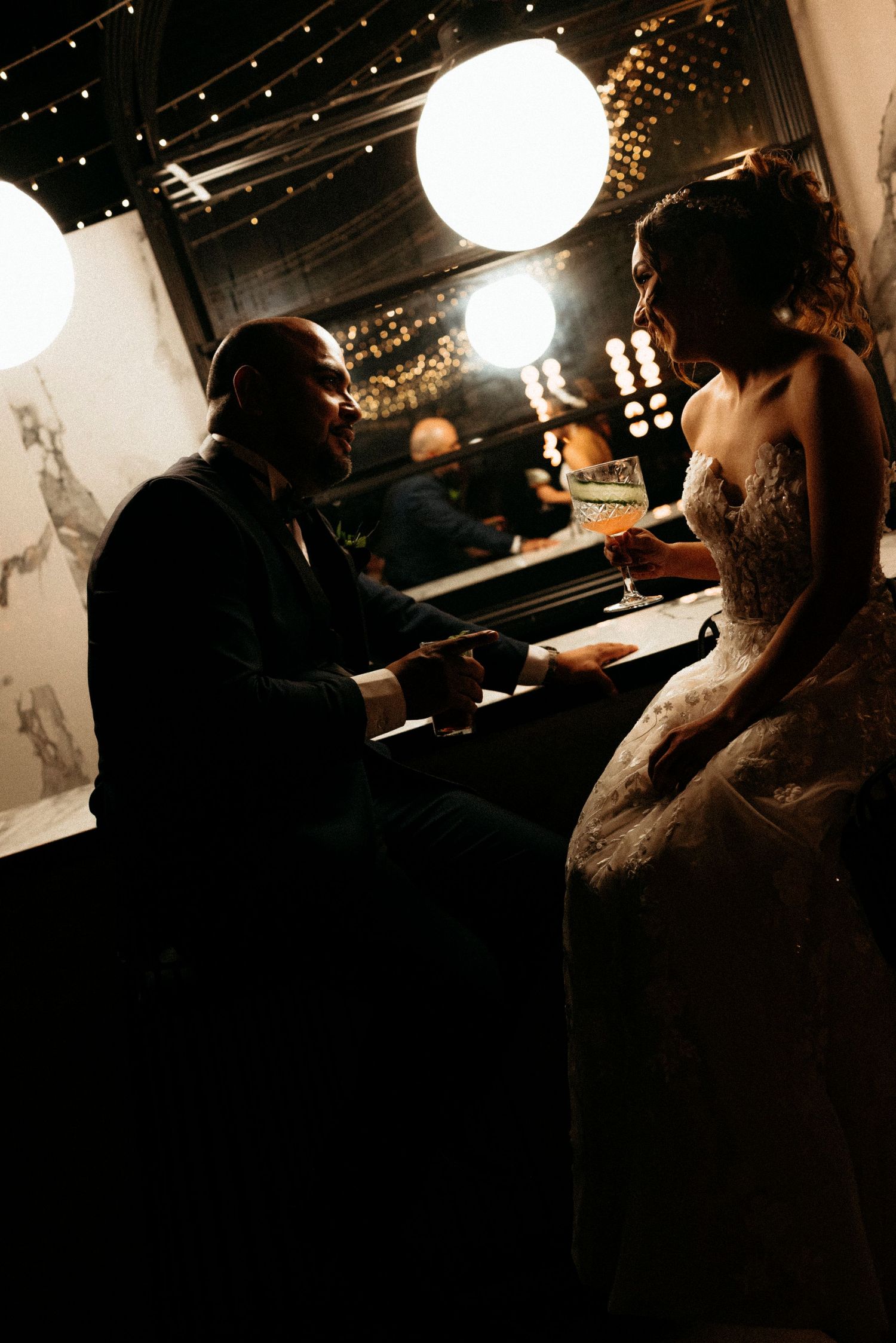 Silhouetted figures sit at a dimly lit bar with glowing round pendant lights overhead creating a moody atmosphere.