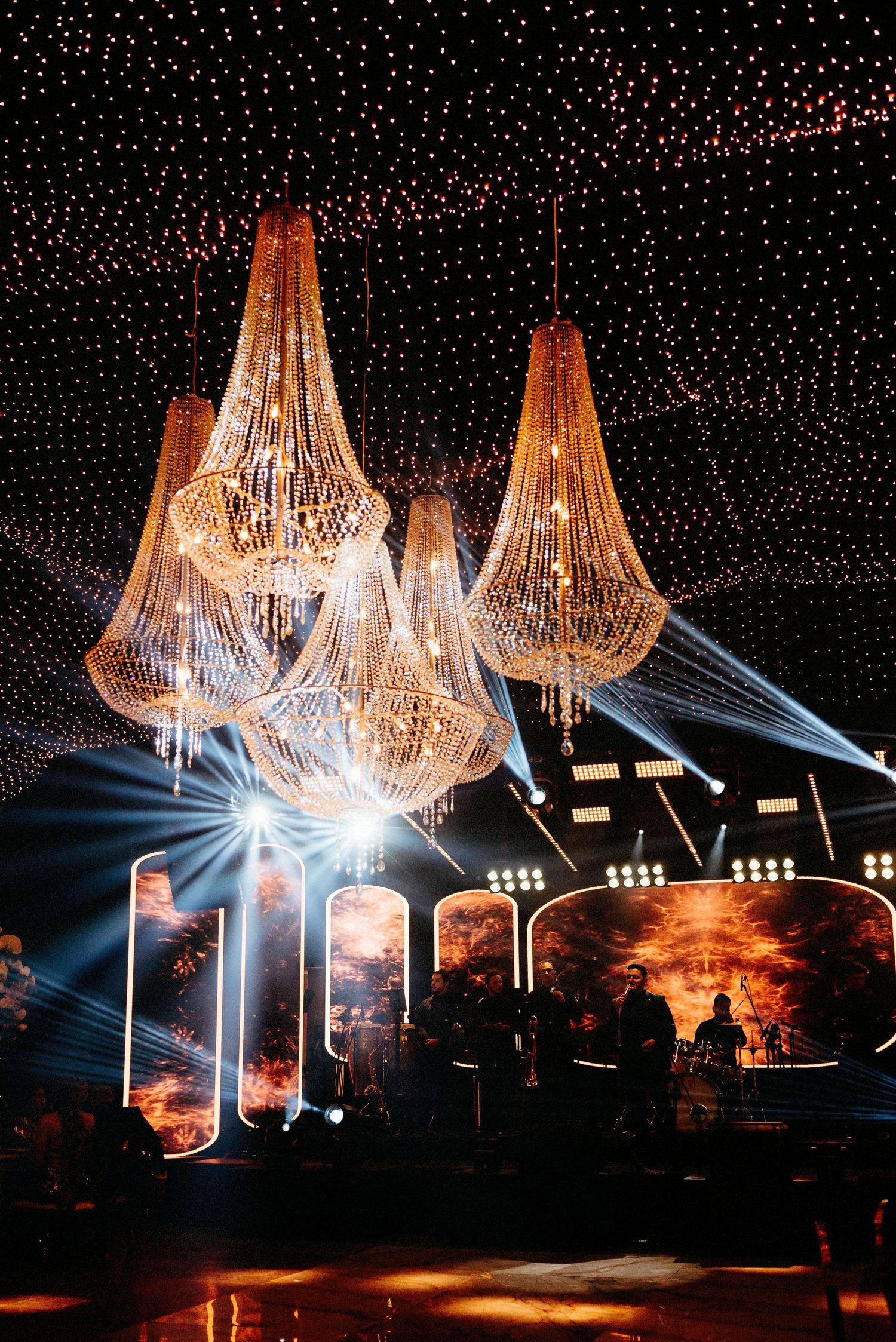 Ornate crystal chandeliers and fairy lights create a magical atmosphere above a stage setup.