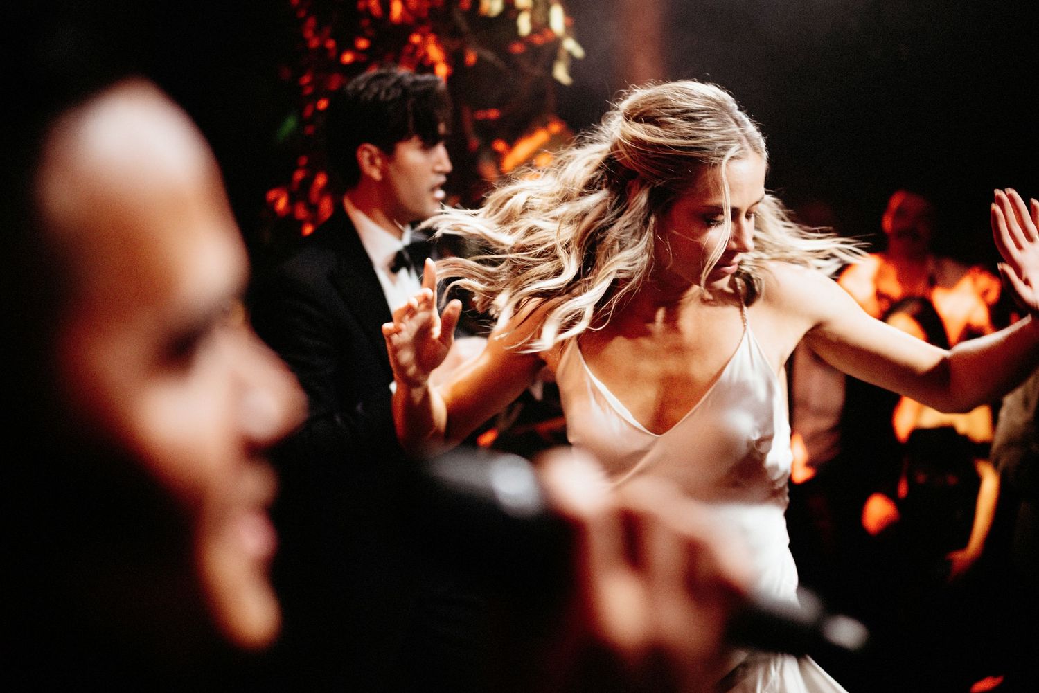 Dancing wedding guests celebrate on a dimly lit dance floor with energetic movements.
