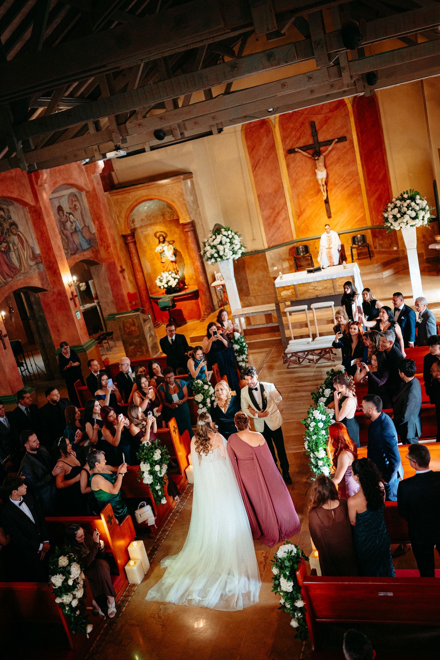 Aerial view of a wedding ceremony in a beautiful church with floral aisle decorations and guests seated in wooden pews.