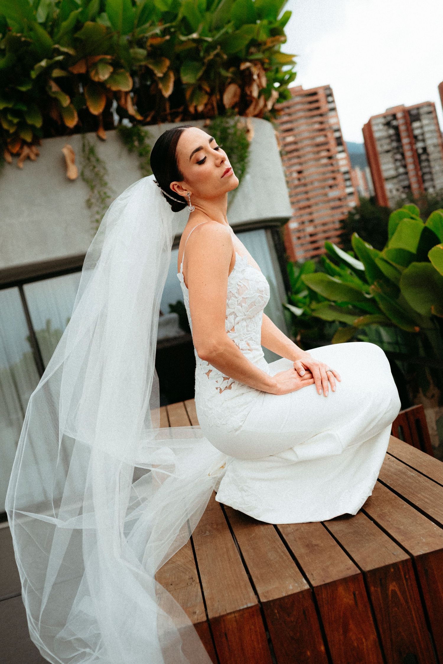 Bride in white lace gown and veil sits on wooden deck with city and greenery backdrop.