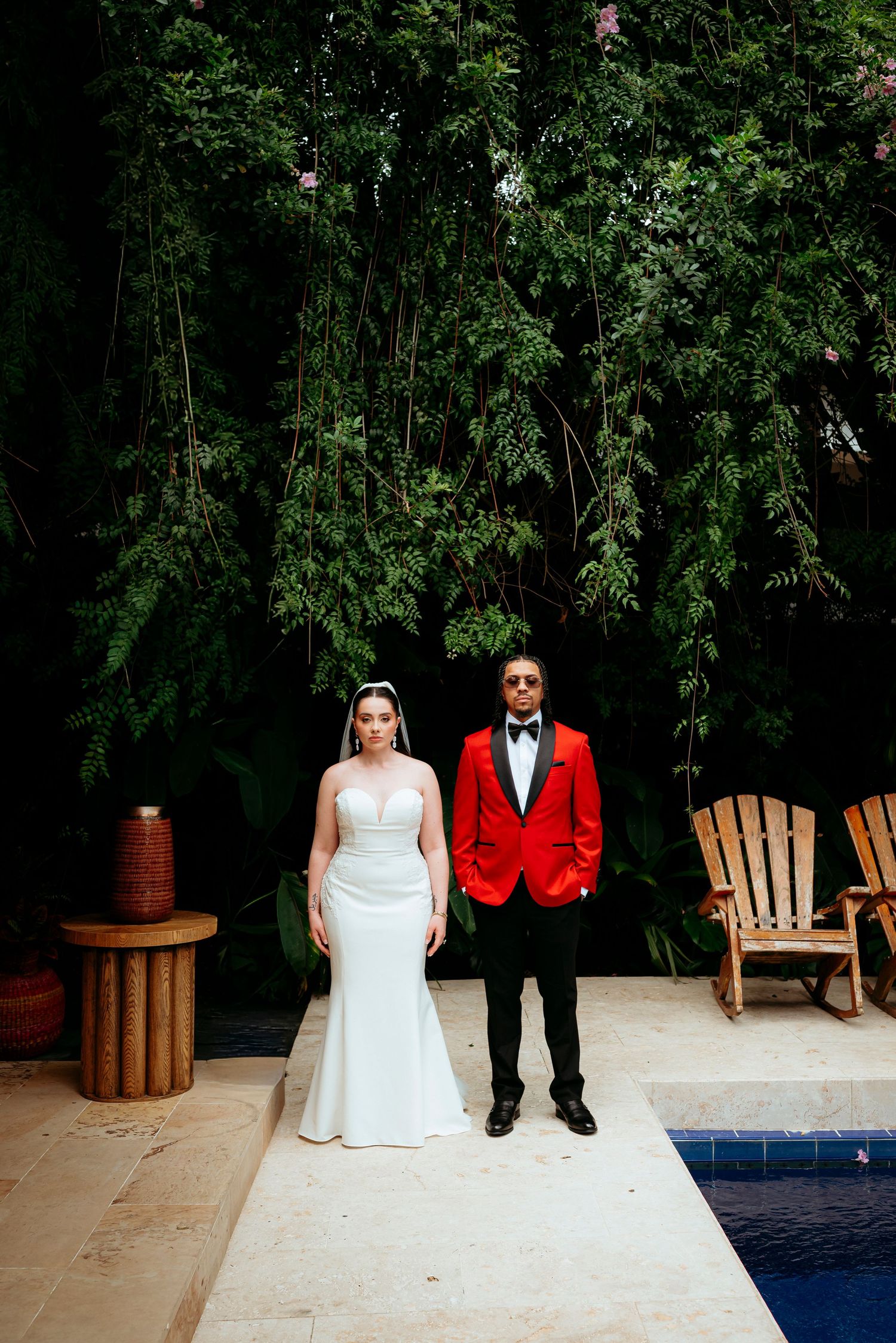 Bride in white gown and groom in red tuxedo jacket stand by a pool surrounded by lush greenery.