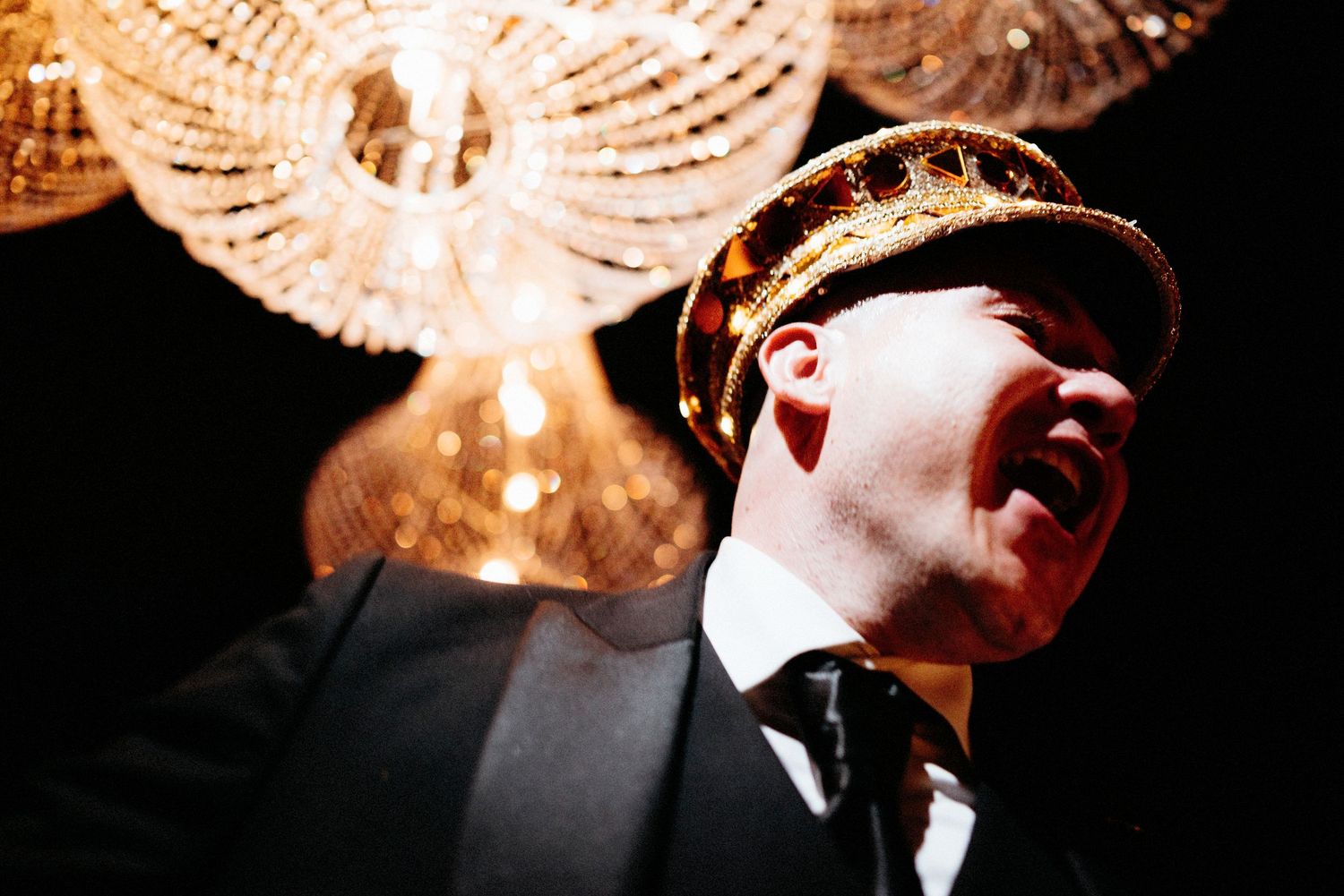A dramatic performer in a suit and cap singing on stage under a crystal chandelier.