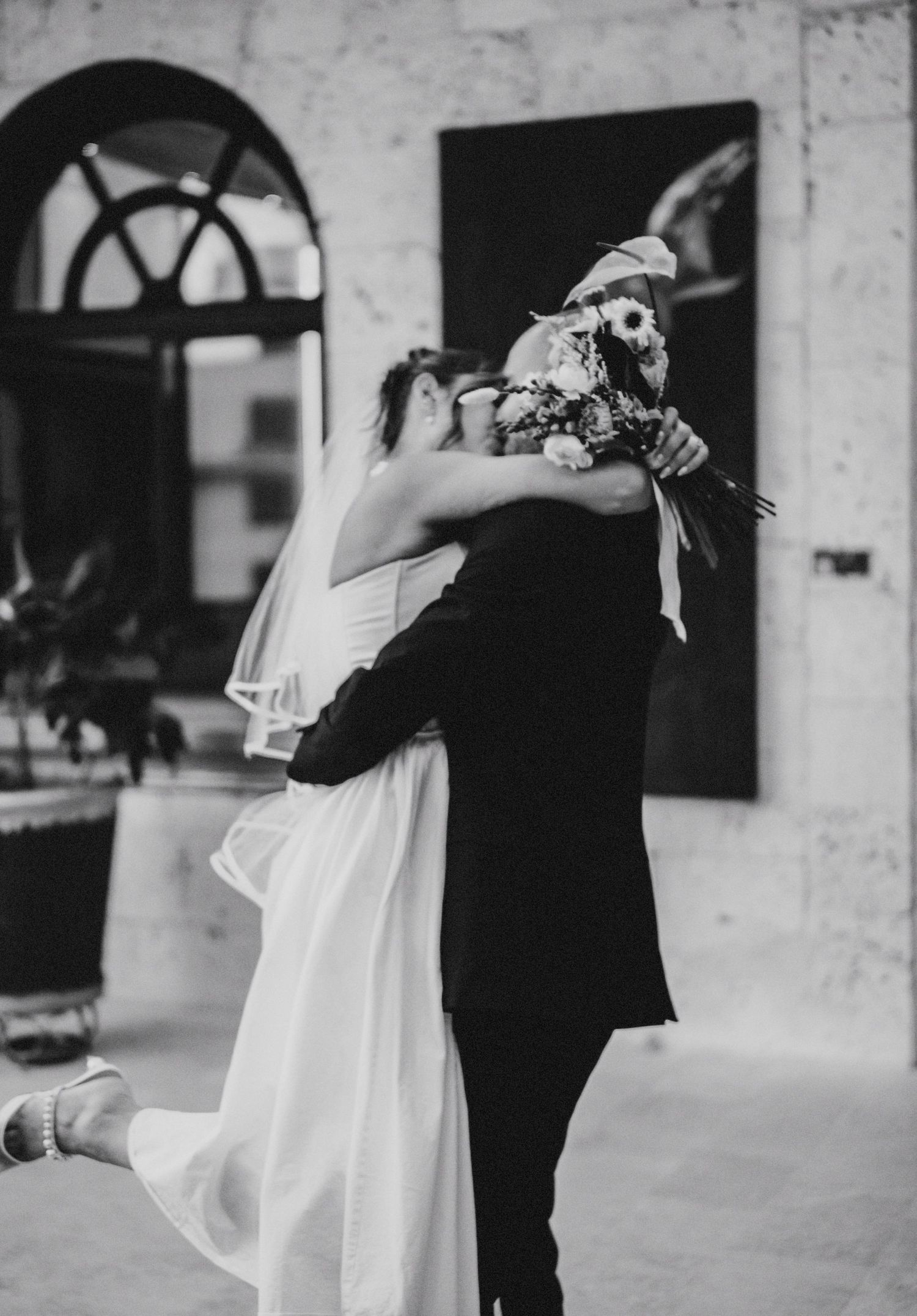 A tender monochromatic moment at a wedding as a couple embraces, with the bride in white and groom in black.