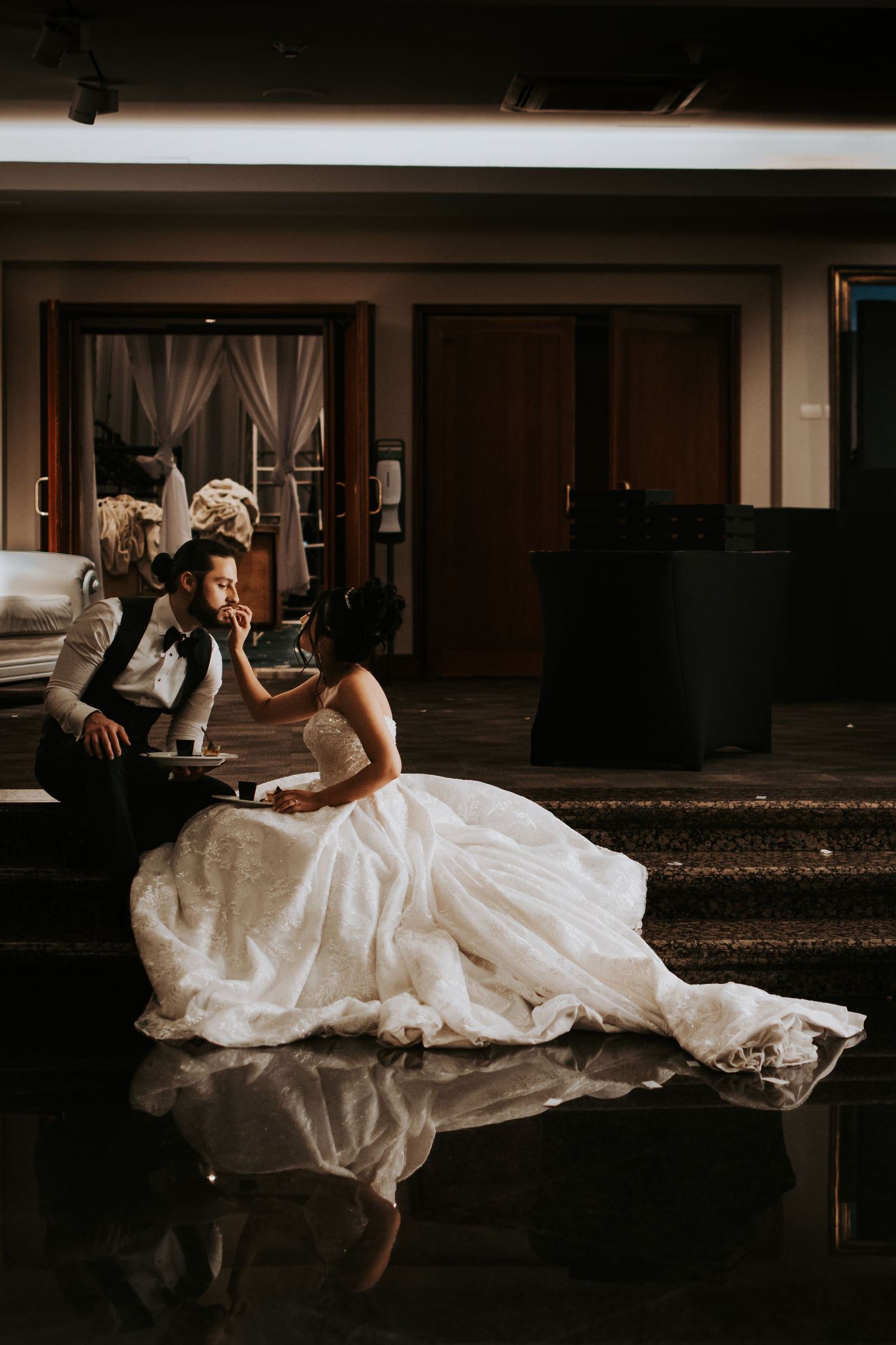 A couple in wedding attire lies on a wooden floor in a dimly lit room creating artistic silhouettes.