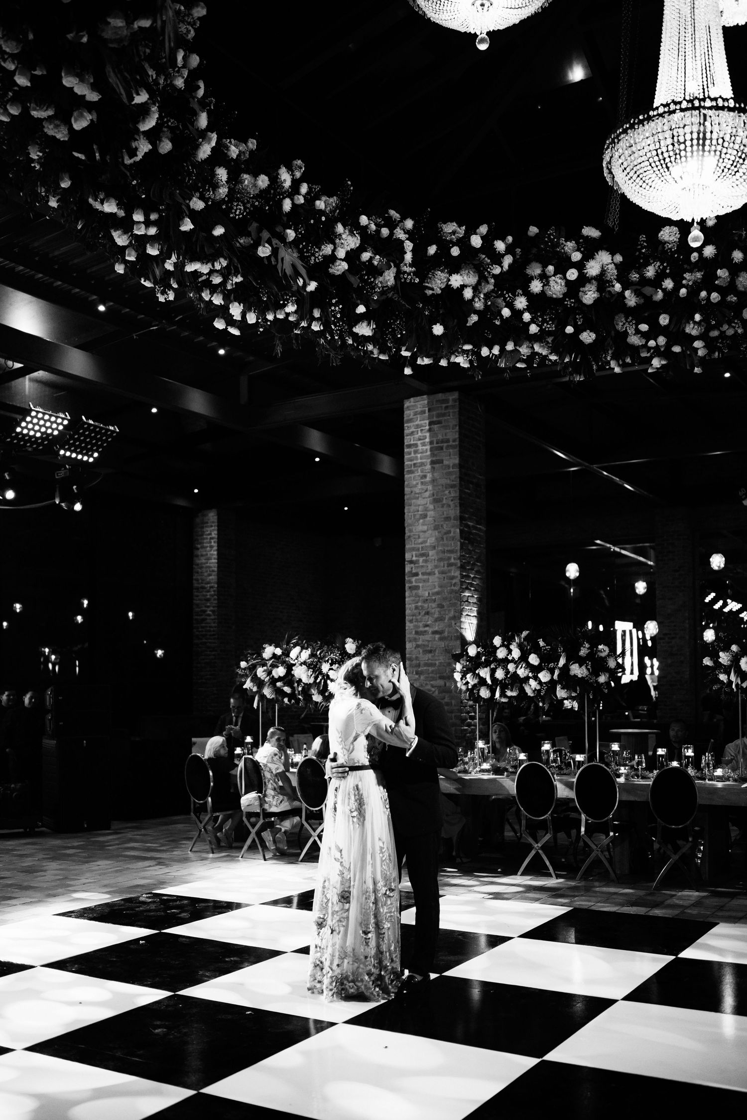 A couple shares their first dance on a black and white checkered floor under elegant chandeliers at an indoor reception.