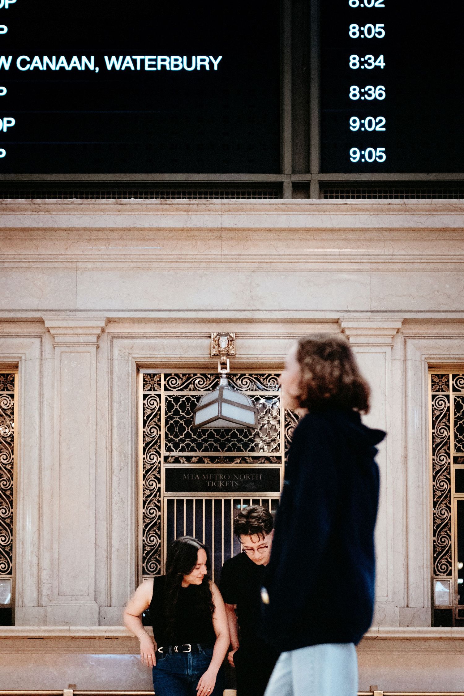 People standing at a train station departure board displaying times for New Canaan and Waterbury.