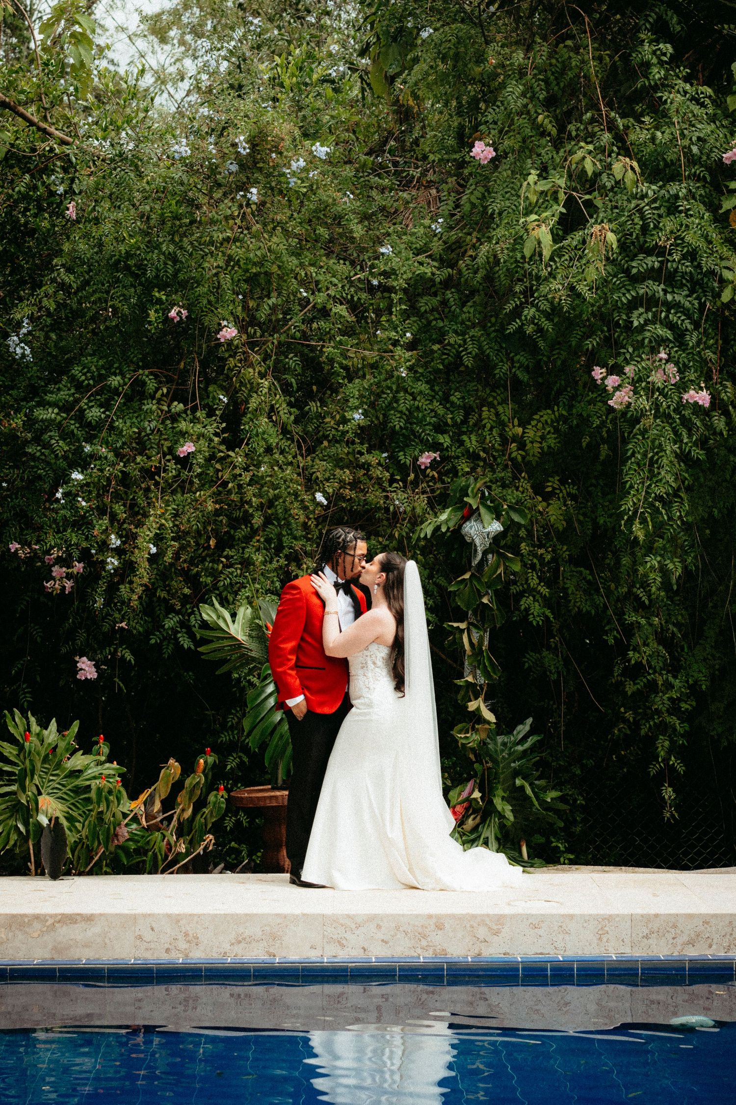 Bride and groom kissing poolside in lush garden, groom in red tuxedo, bride in white gown with veil.