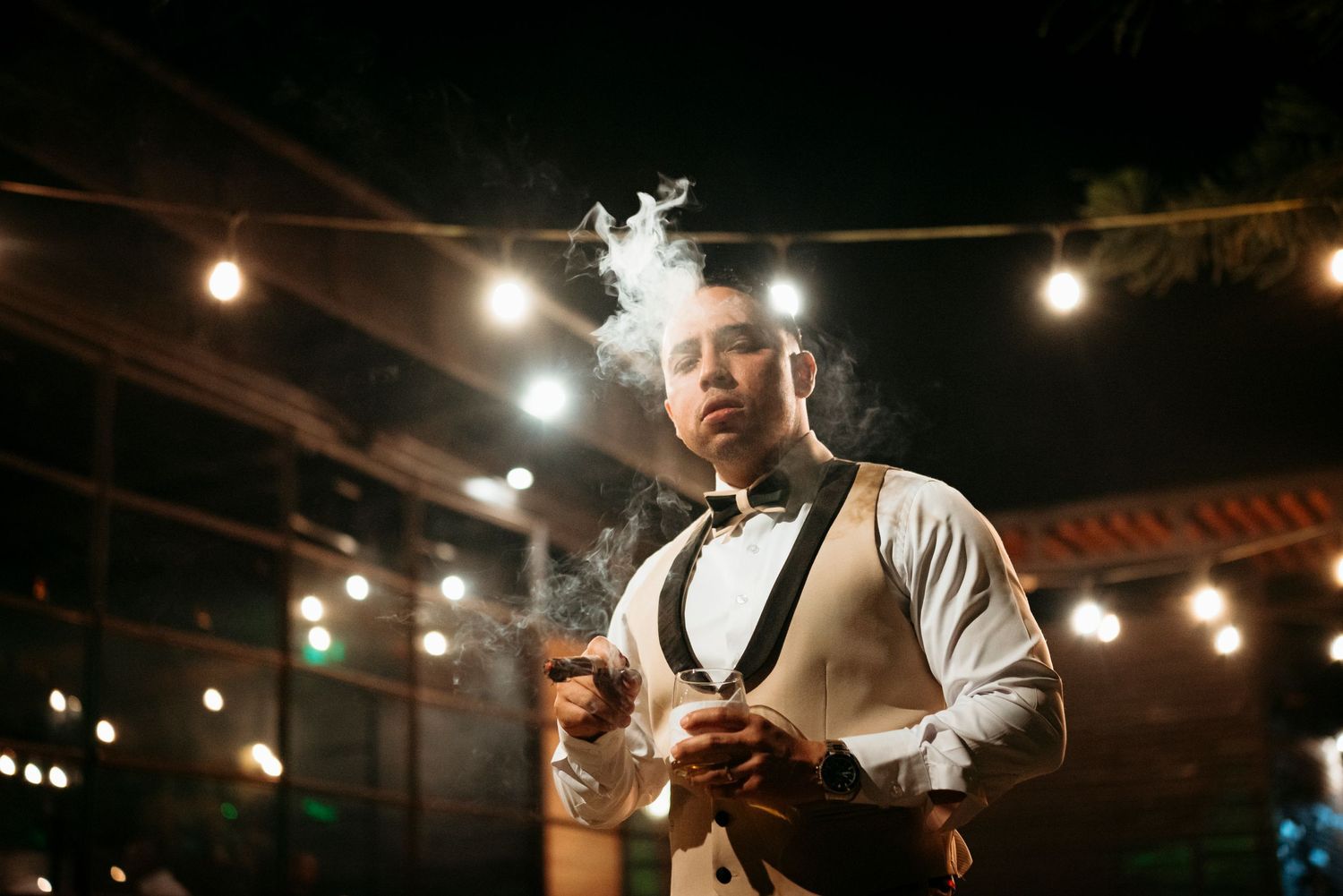 An elegant server in bow tie and vest holding drinks under warm string lights at an evening event.