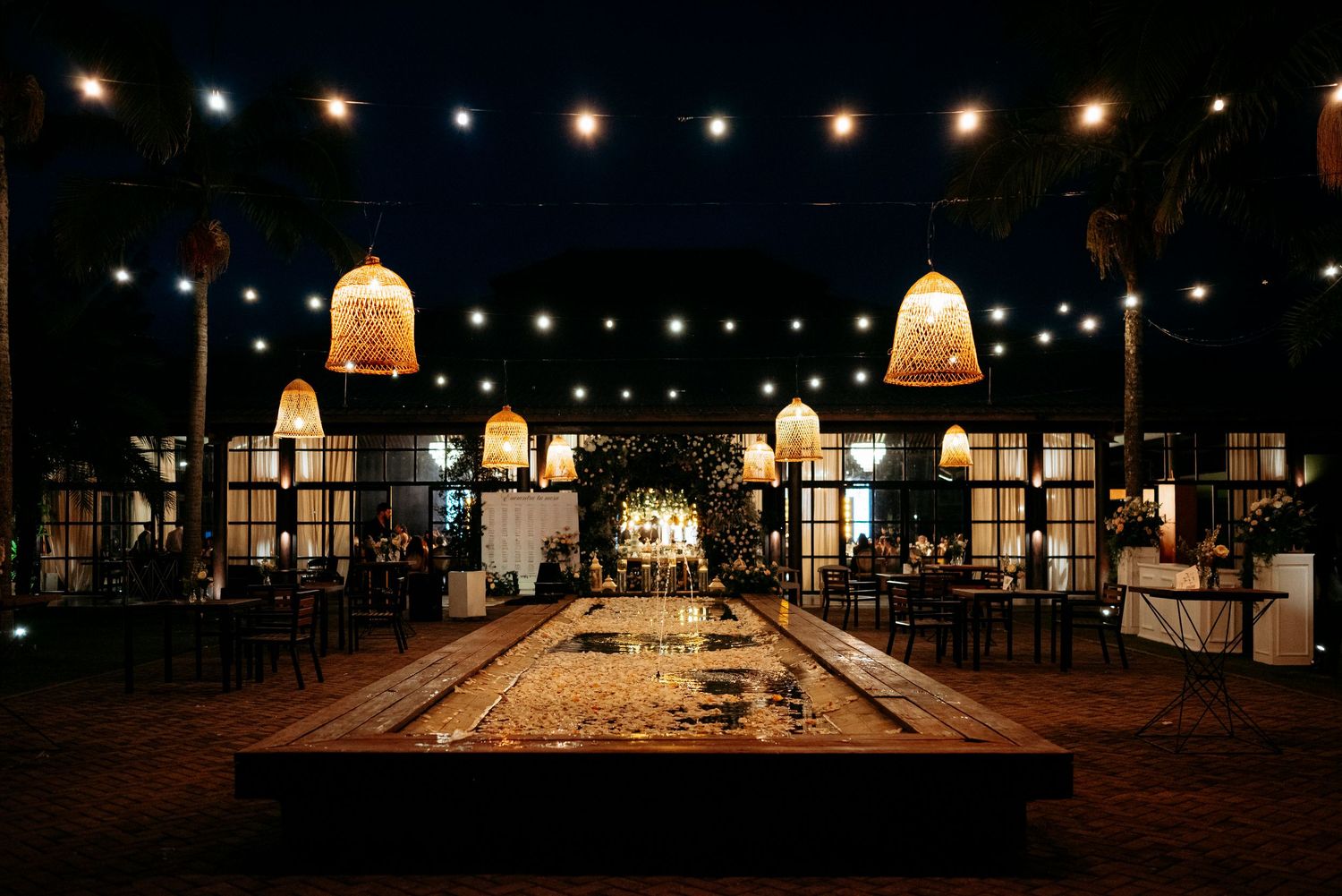 Elegant outdoor venue at night with string lights, wicker pendant lamps, and a reflective water feature centerpiece.