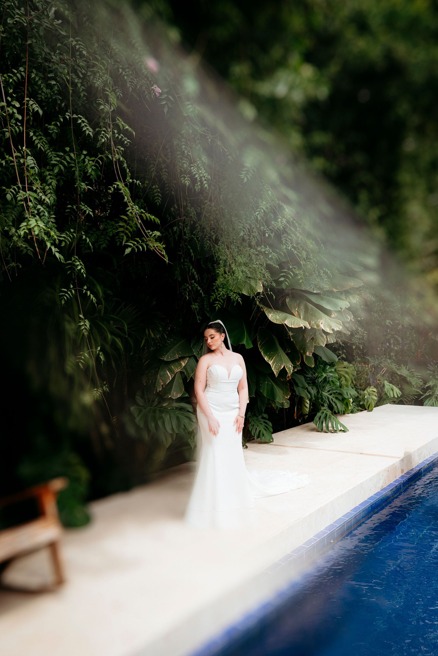 Bride in white gown standing by a tropical pool surrounded by lush green foliage in soft natural light.