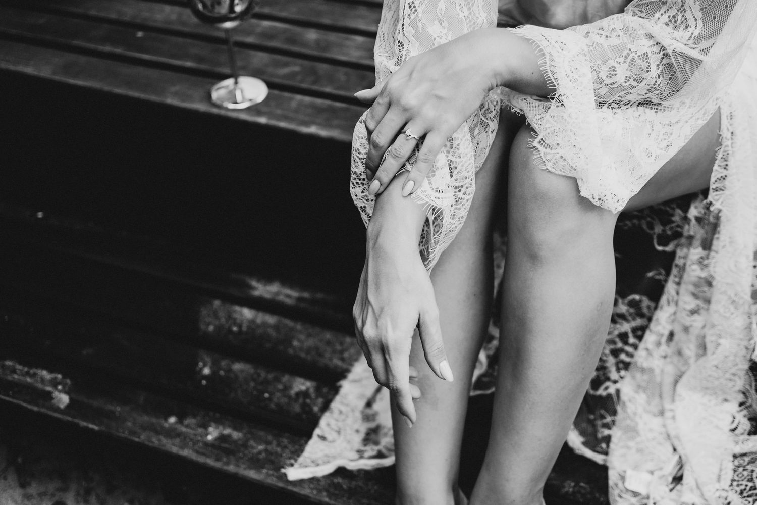 Black and white photo of a bride showing delicate lace garter on leg while sitting on wooden steps.