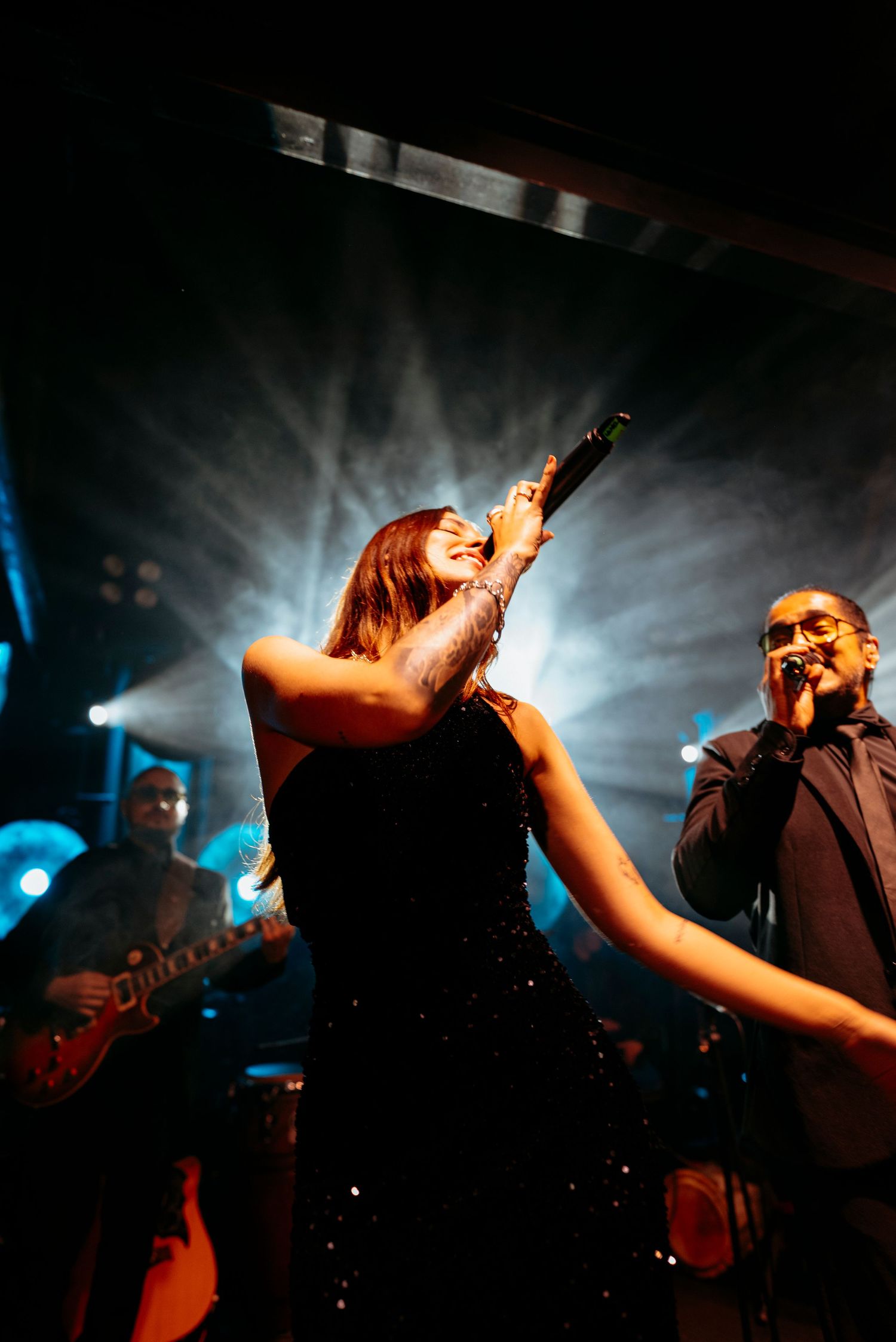 Female vocalist holds microphone aloft on stage with dramatic light beams, alongside male singer and guitarist in dark venue.