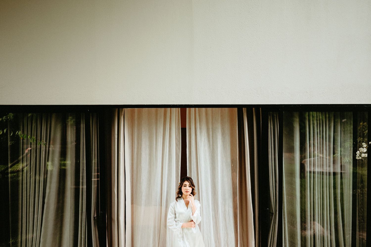 A person in a white dress stands between dark and light curtains in an elegant interior space.