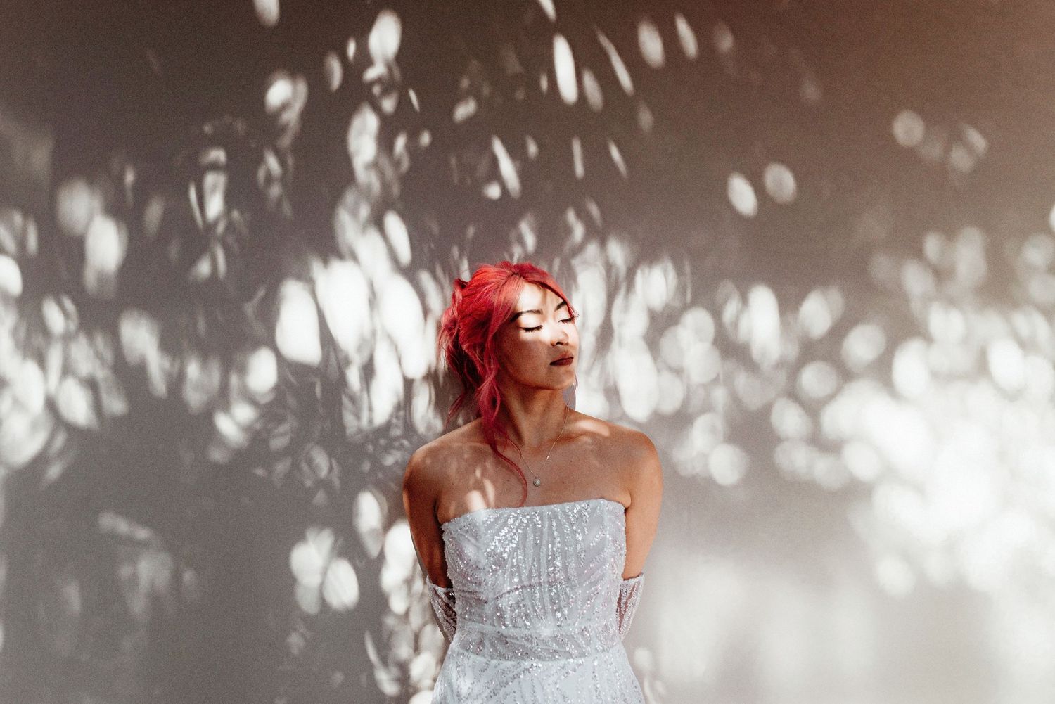 A person with vibrant red hair wearing a white lace dress poses in dappled sunlight against a natural background.