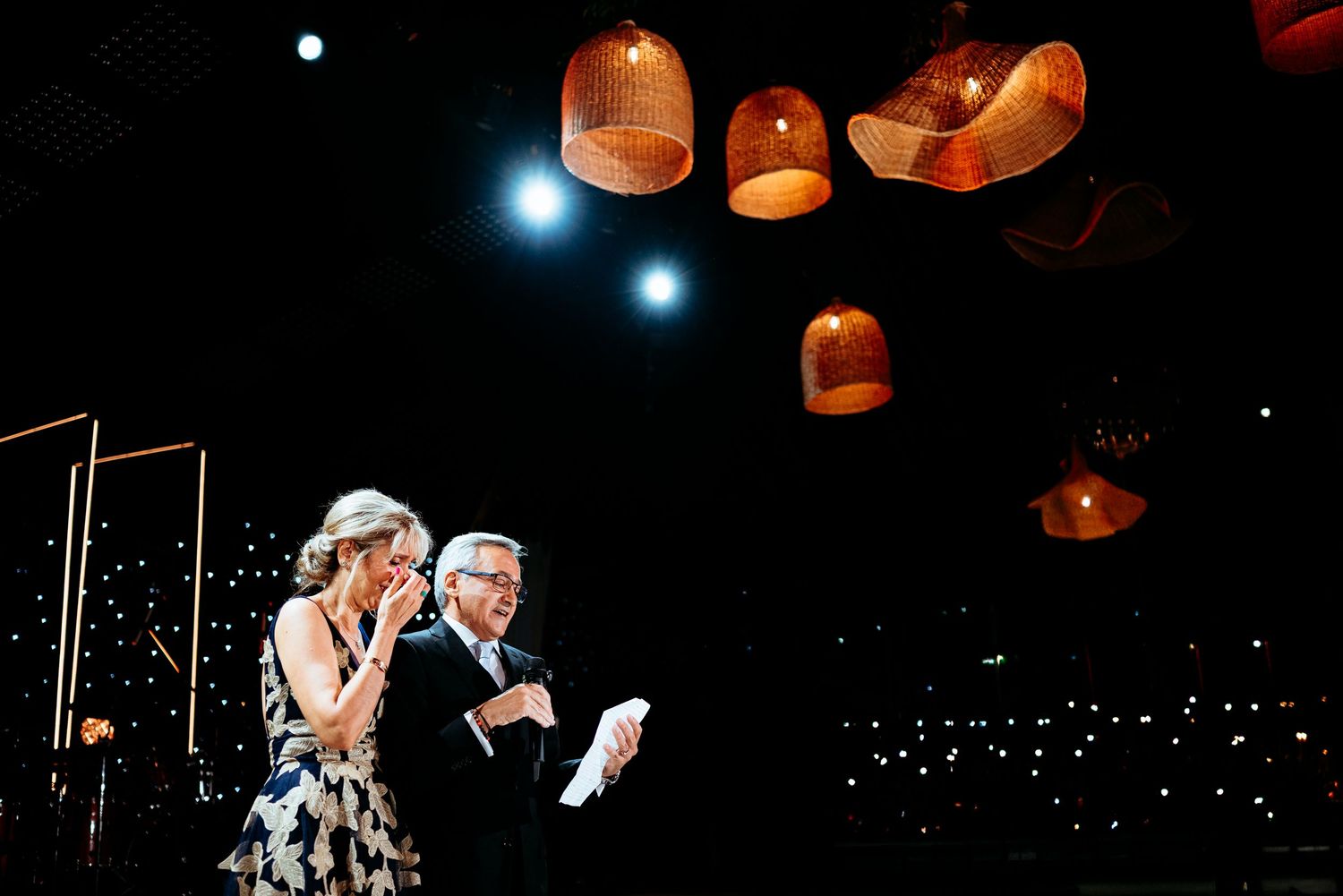 Two elegantly dressed speakers present on a darkened stage beneath warm glowing wicker pendant lights.