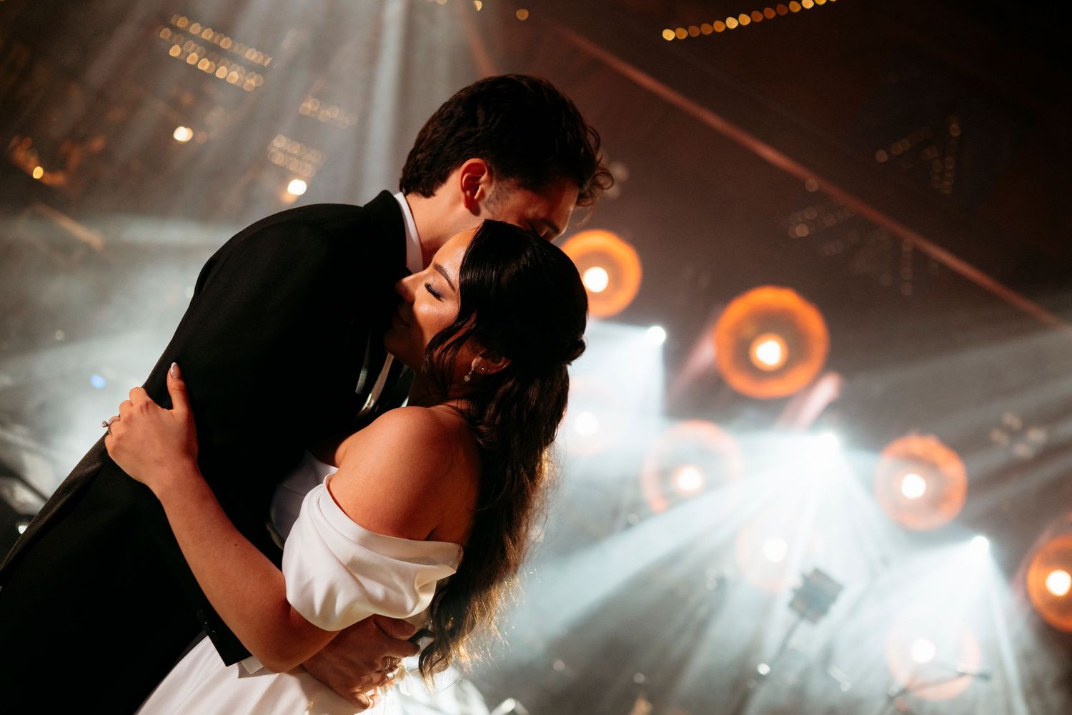Bride and groom sharing a romantic first dance kiss under dramatic stage lights at wedding reception.