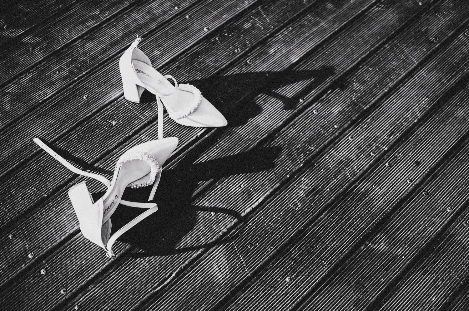Pair of elegant white high heel shoes laying on a dark wooden floor casting dramatic shadows in black and white.