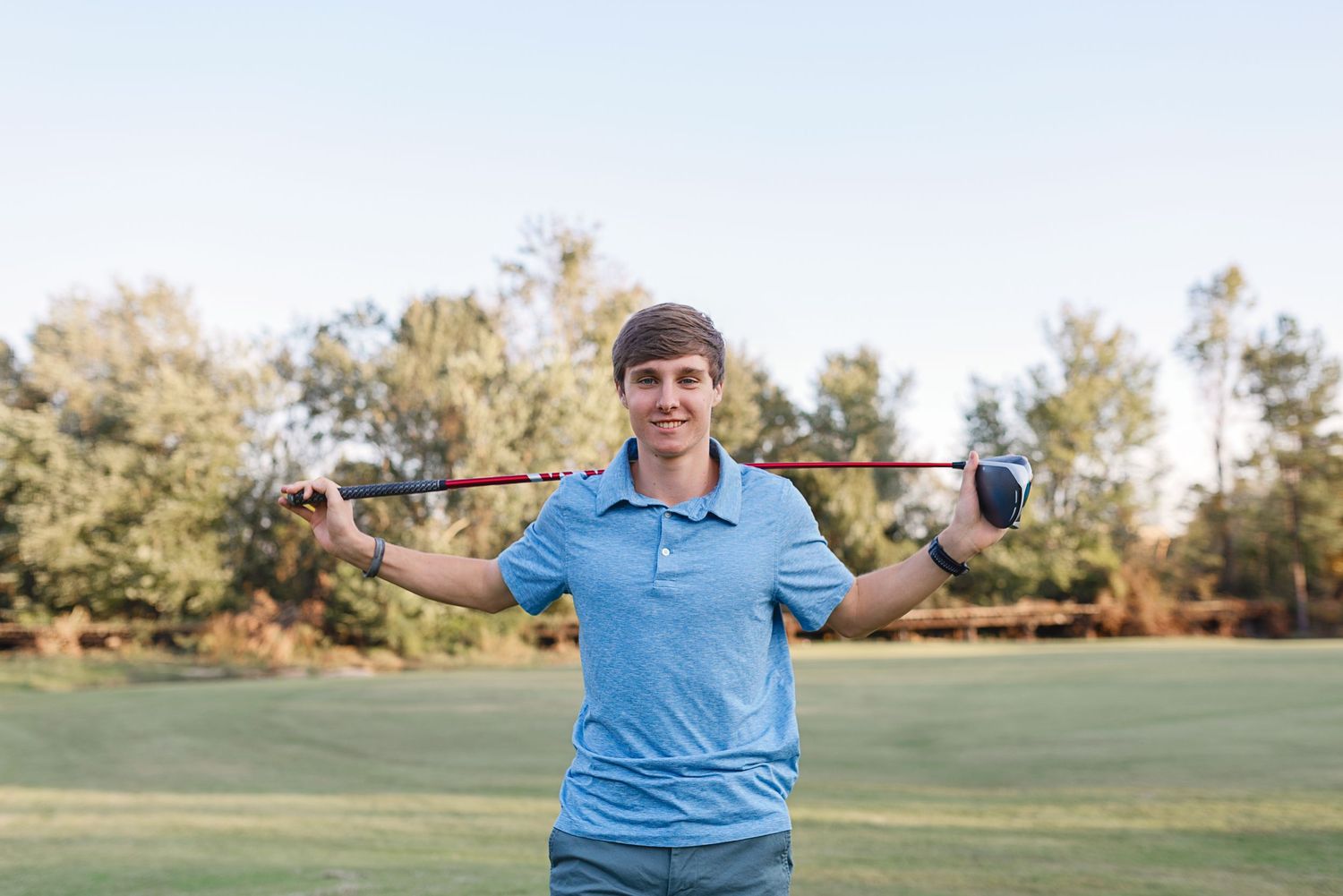 Golfer in light blue polo shirt holding club across shoulders on green course.