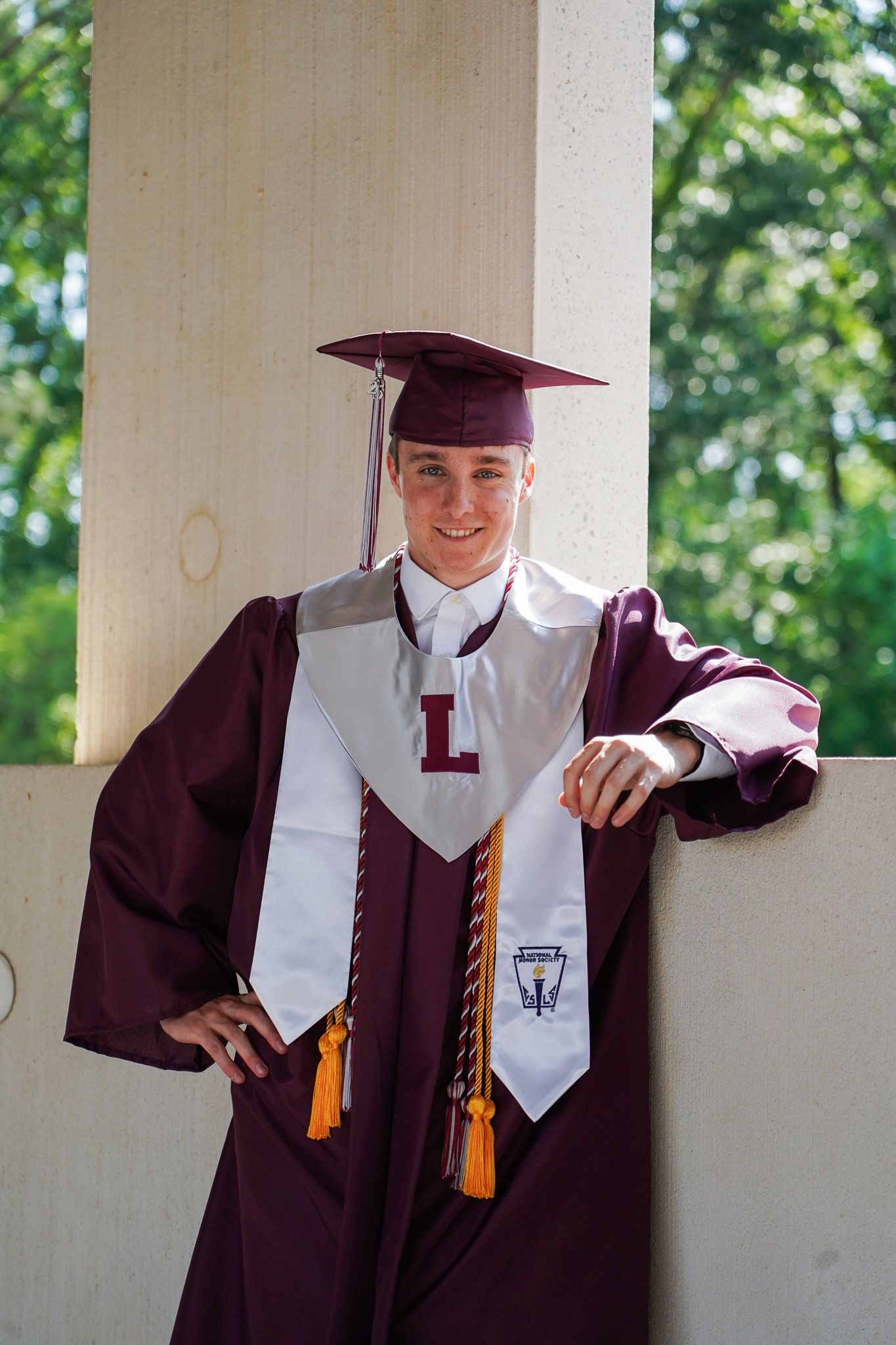 Graduate in maroon cap and gown poses against white columns during graduation photoshoot.
