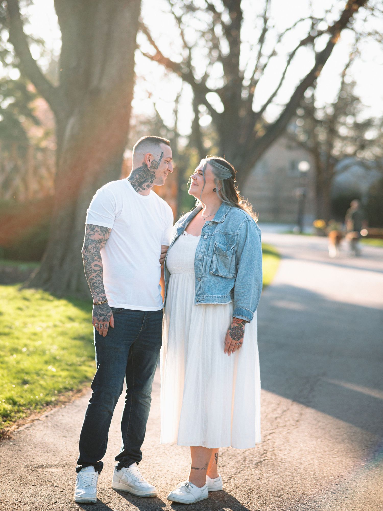 Couple smiling at each other outdoors, man with tattoos in white tee, woman in white dress and denim jacket, sunny park.