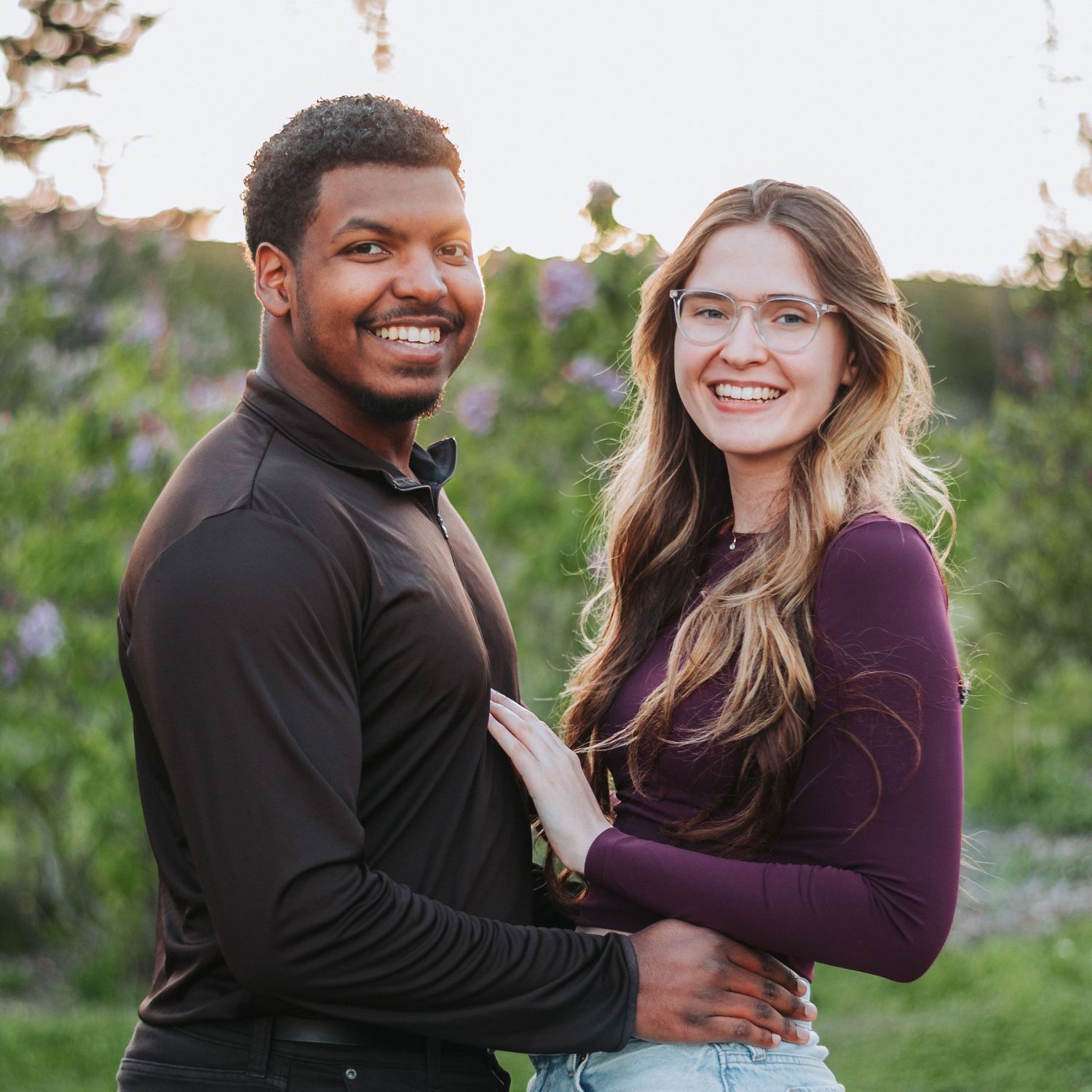 Smiling interracial couple embracing outdoors at sunset with wildflowers in background.