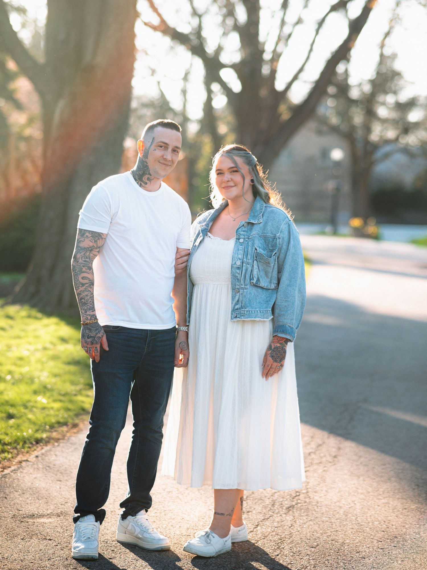 A tattooed couple poses on a sunlit path, man in white tee and jeans, woman in white dress and denim jacket.