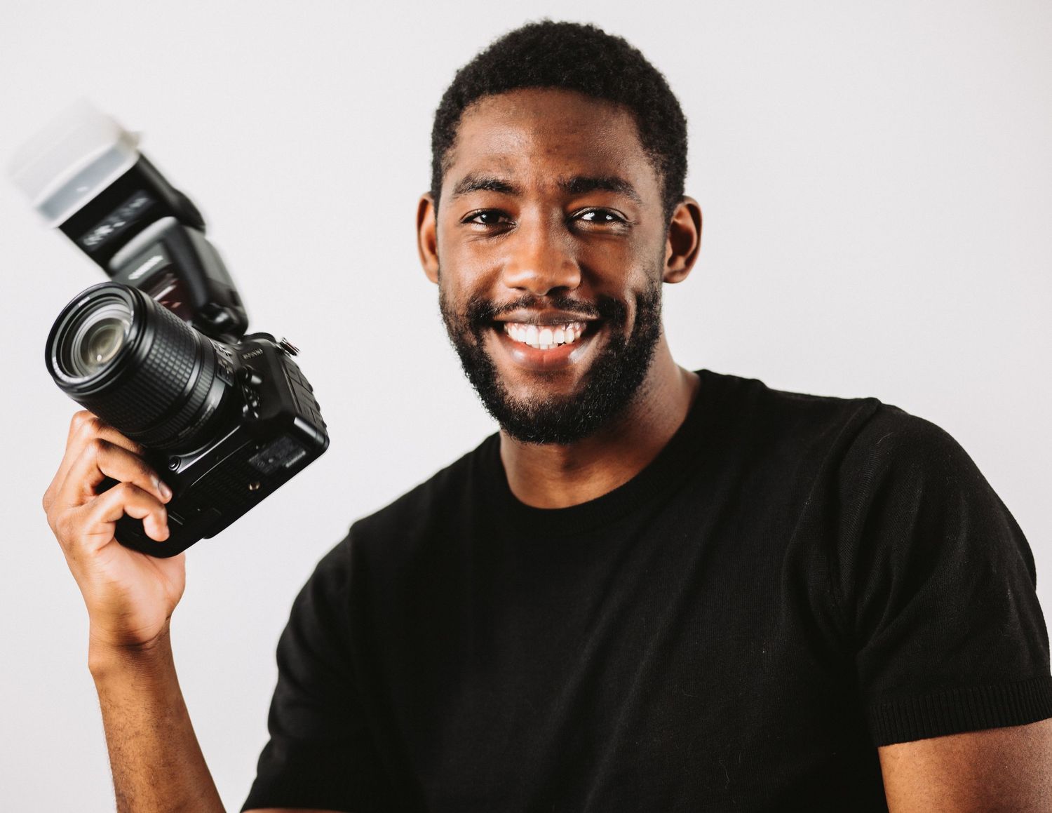 Smiling young man holding a DSLR camera with flash in a studio, wearing a black t-shirt on white background.