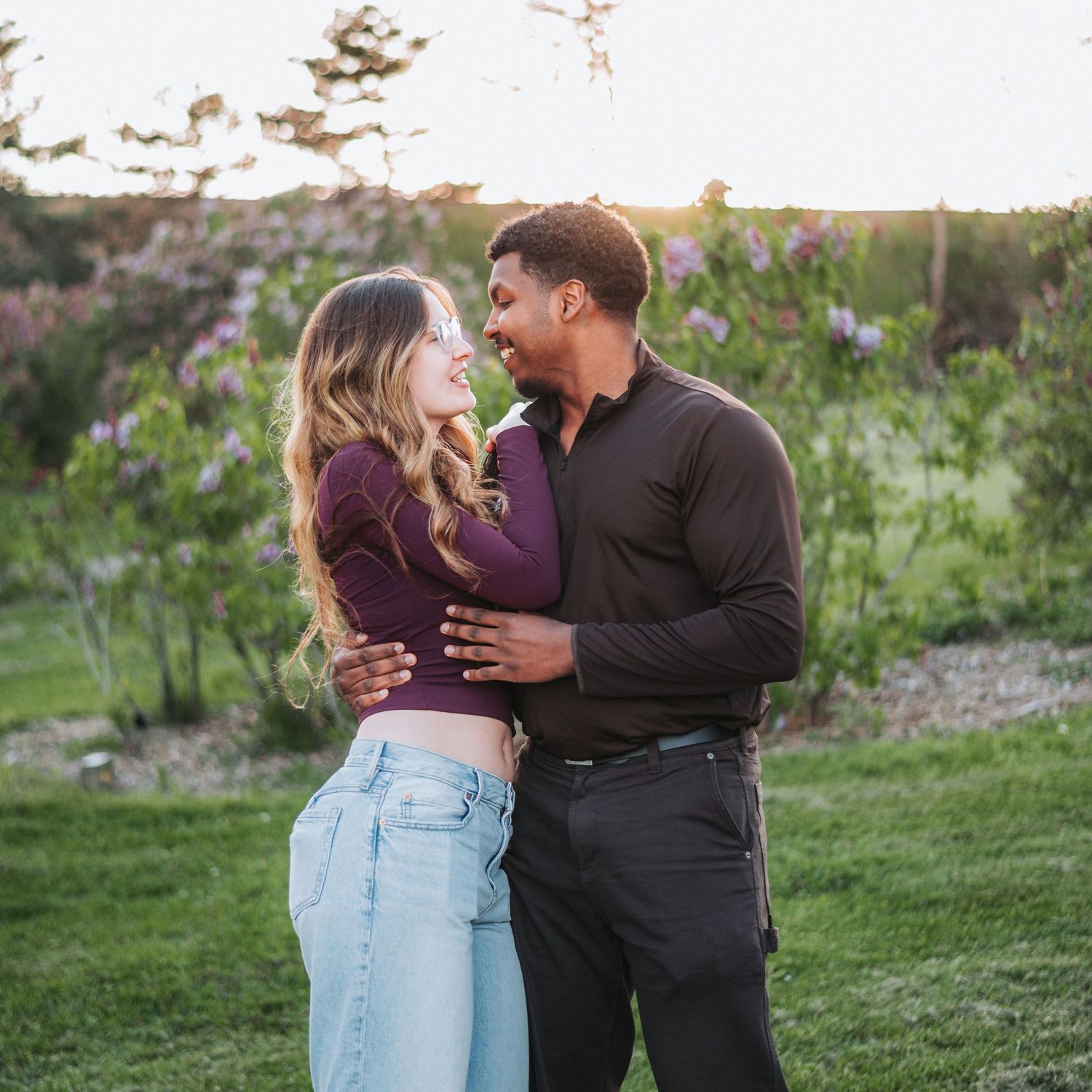 Couple sharing a romantic moment outdoors at sunset, embracing in a lush green garden with blooming flowers.