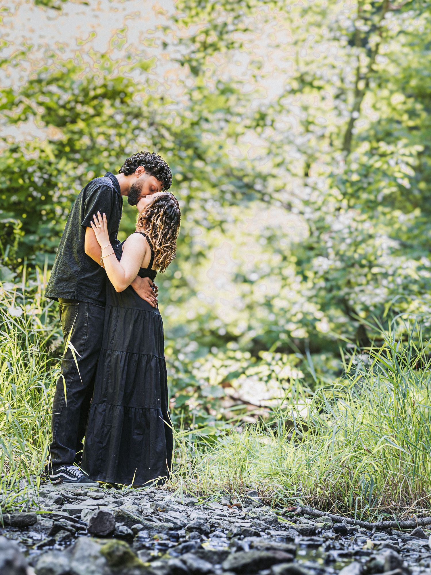 Couple sharing a romantic kiss surrounded by lush green forest and rocky ground in nature.