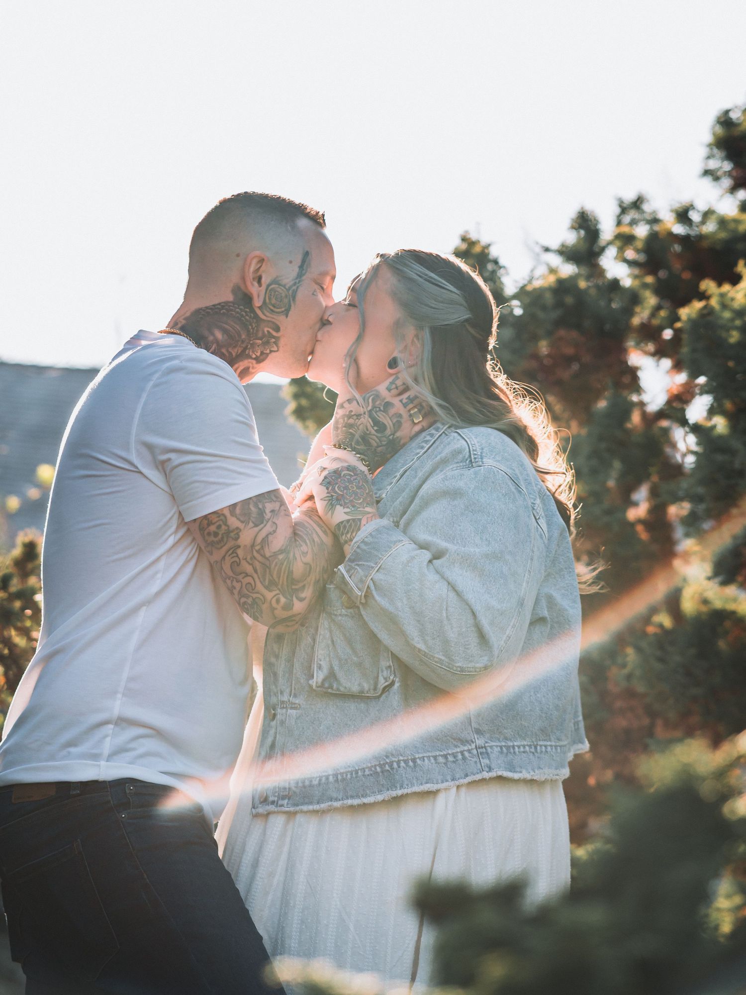 Tattooed couple sharing a romantic kiss outdoors in golden sunlight with trees in the background.