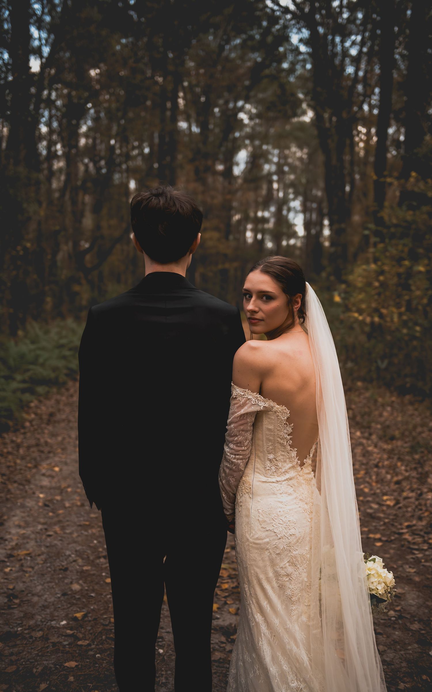 A bride in a lace wedding dress and long veil stands next to a groom in black suit in an autumn forest setting.