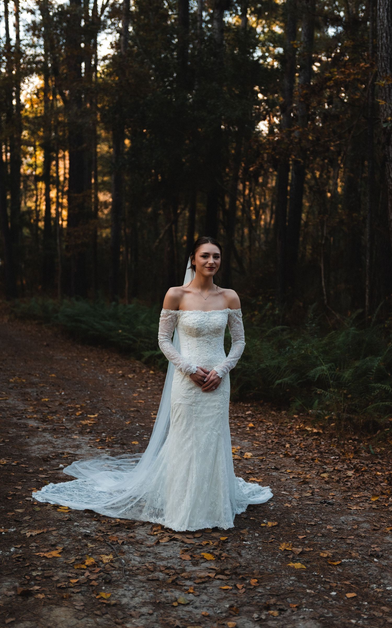 A bride in a white off-shoulder lace wedding dress poses on a forest path during golden hour sunset.