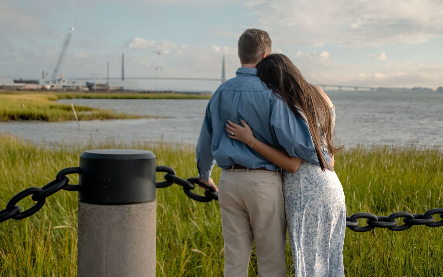 A couple stands together on a coastal boardwalk overlooking a scenic marsh landscape at sunset.