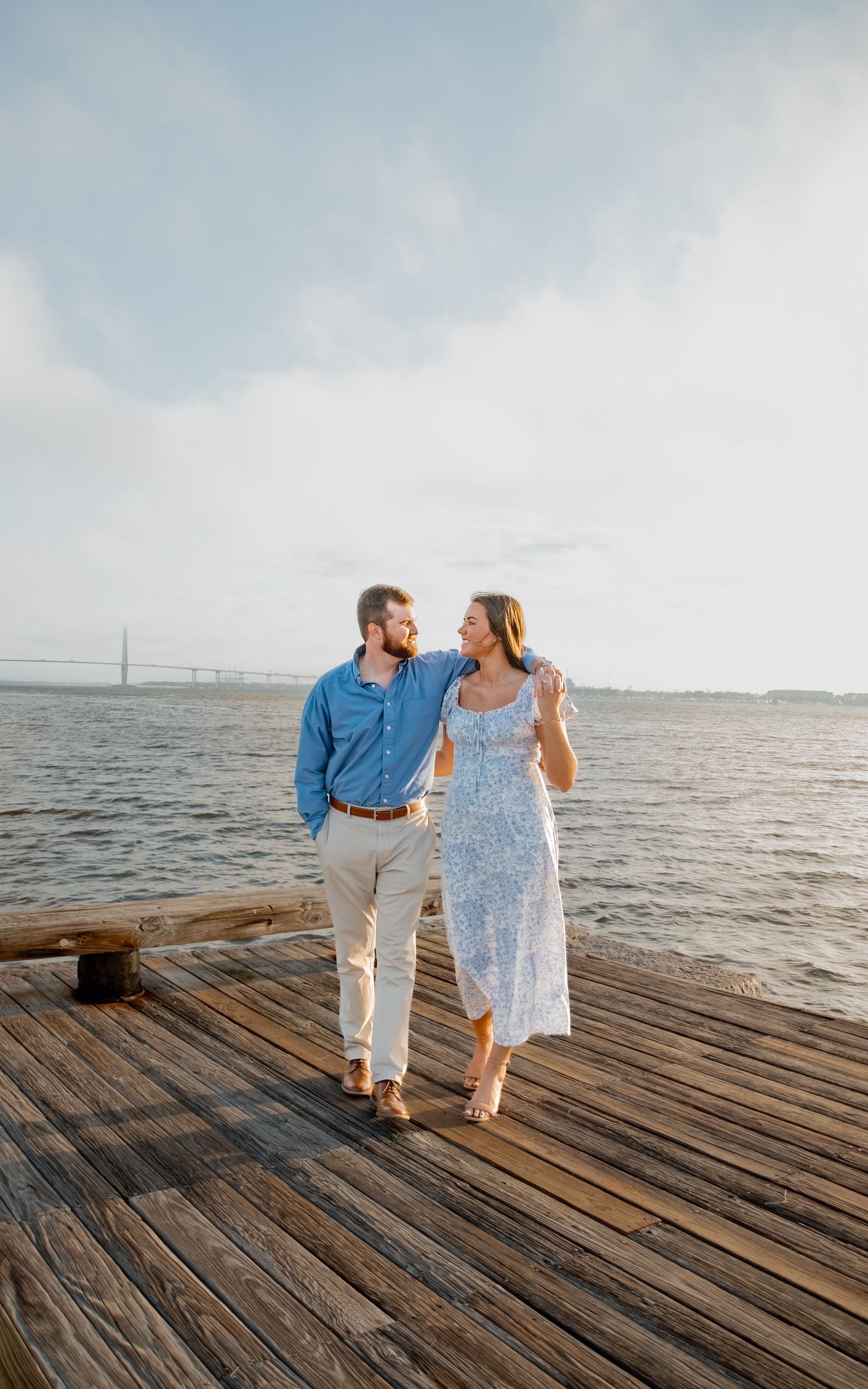 A couple in coordinating blue and white outfits share a romantic moment on a wooden pier by the ocean.