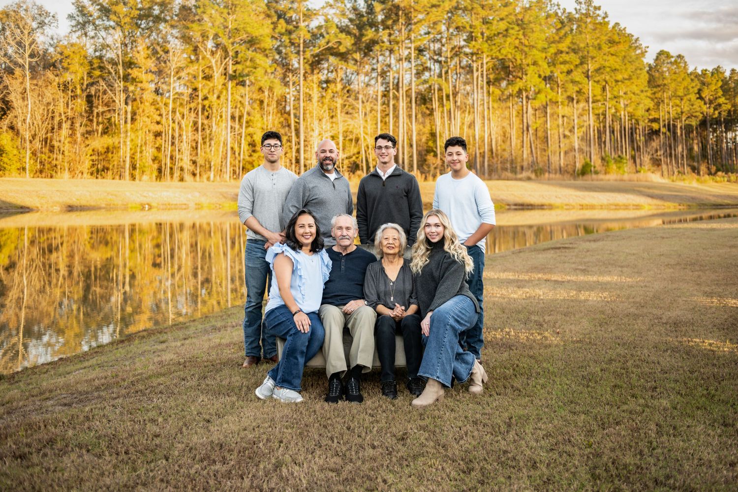 A group sits together on a grassy bank beside a reflective lake with golden sunlit trees in the background.
