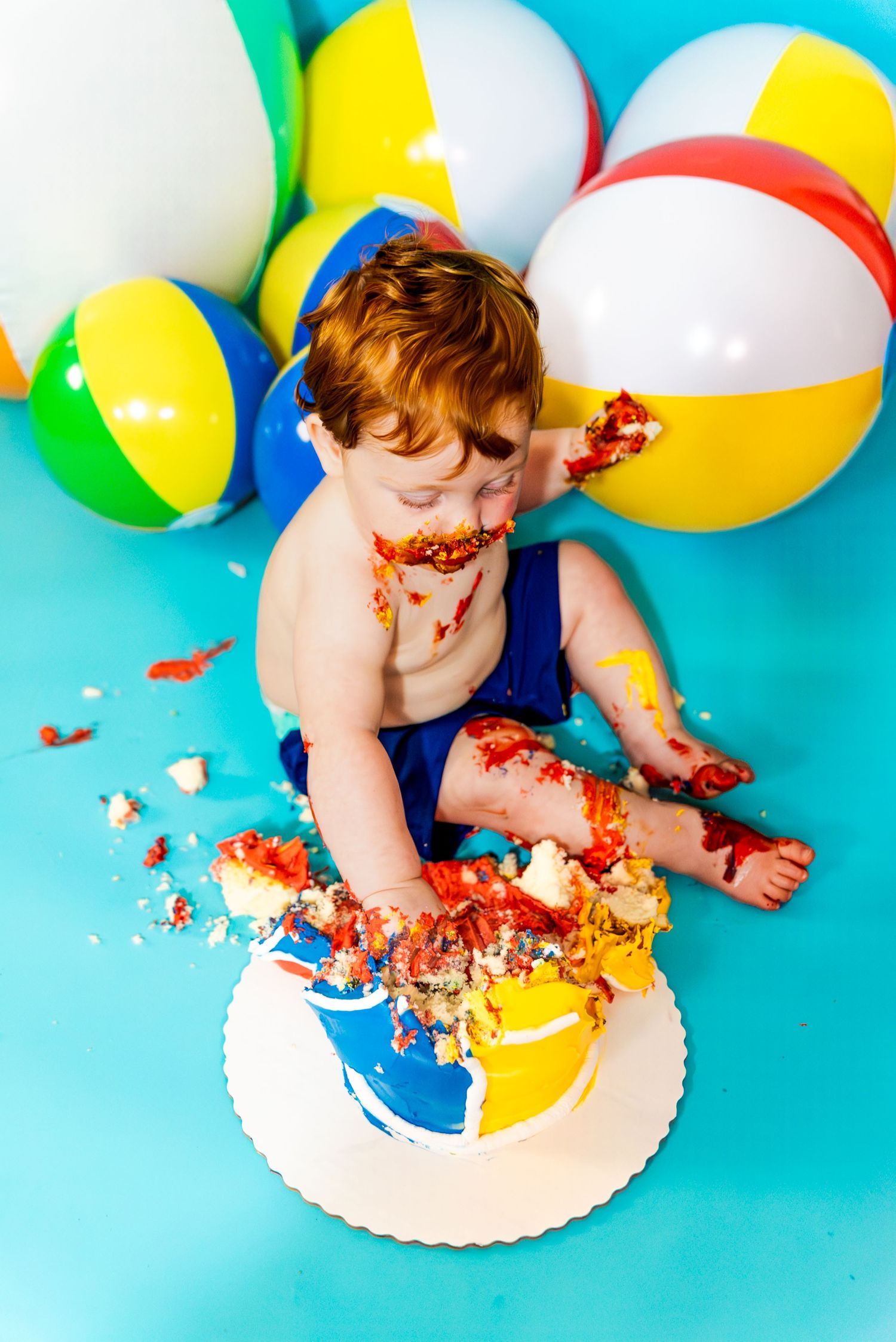 A baby in blue shorts plays with colorful beach balls and a smashed cake against a turquoise backdrop.