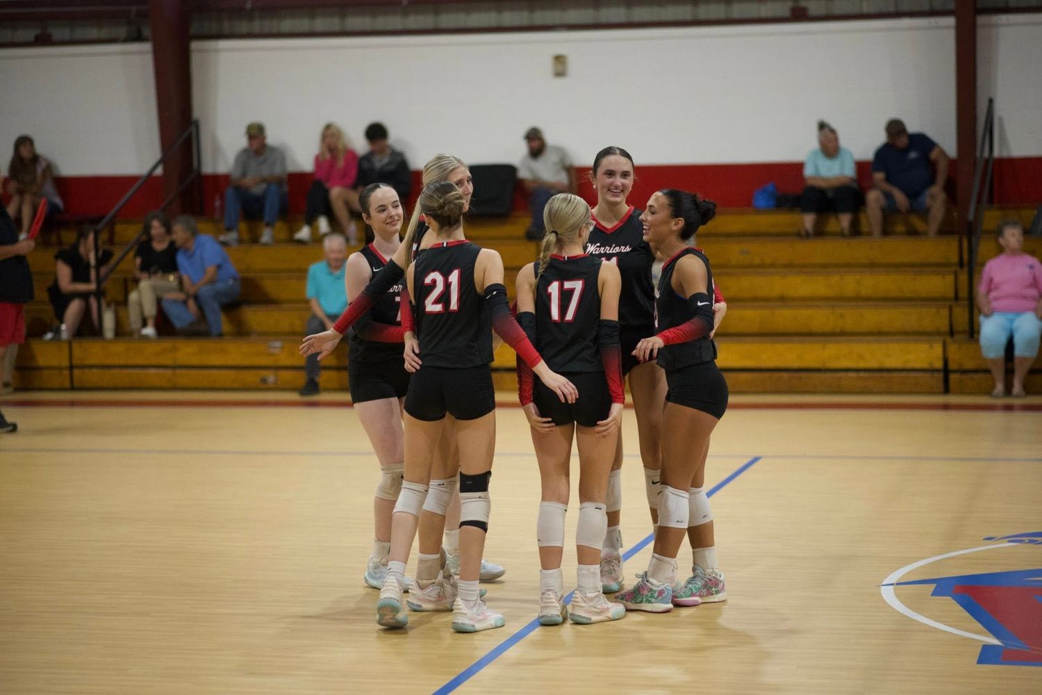 Indoor volleyball players gather around their coach during a timeout in a high school gymnasium.