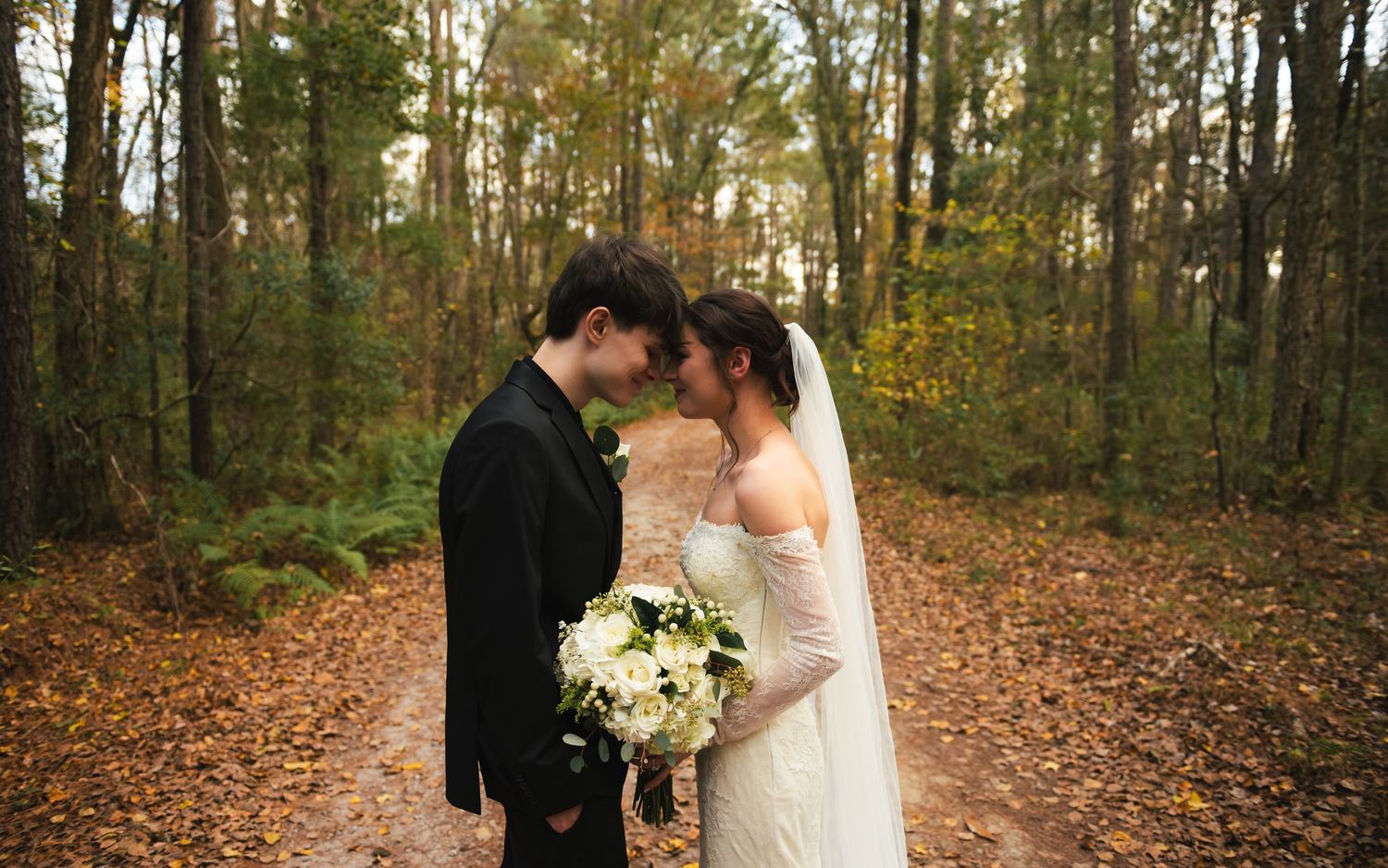 Couple sharing romantic moments in an autumn forest with fallen leaves and tall trees during a wedding photoshoot.
