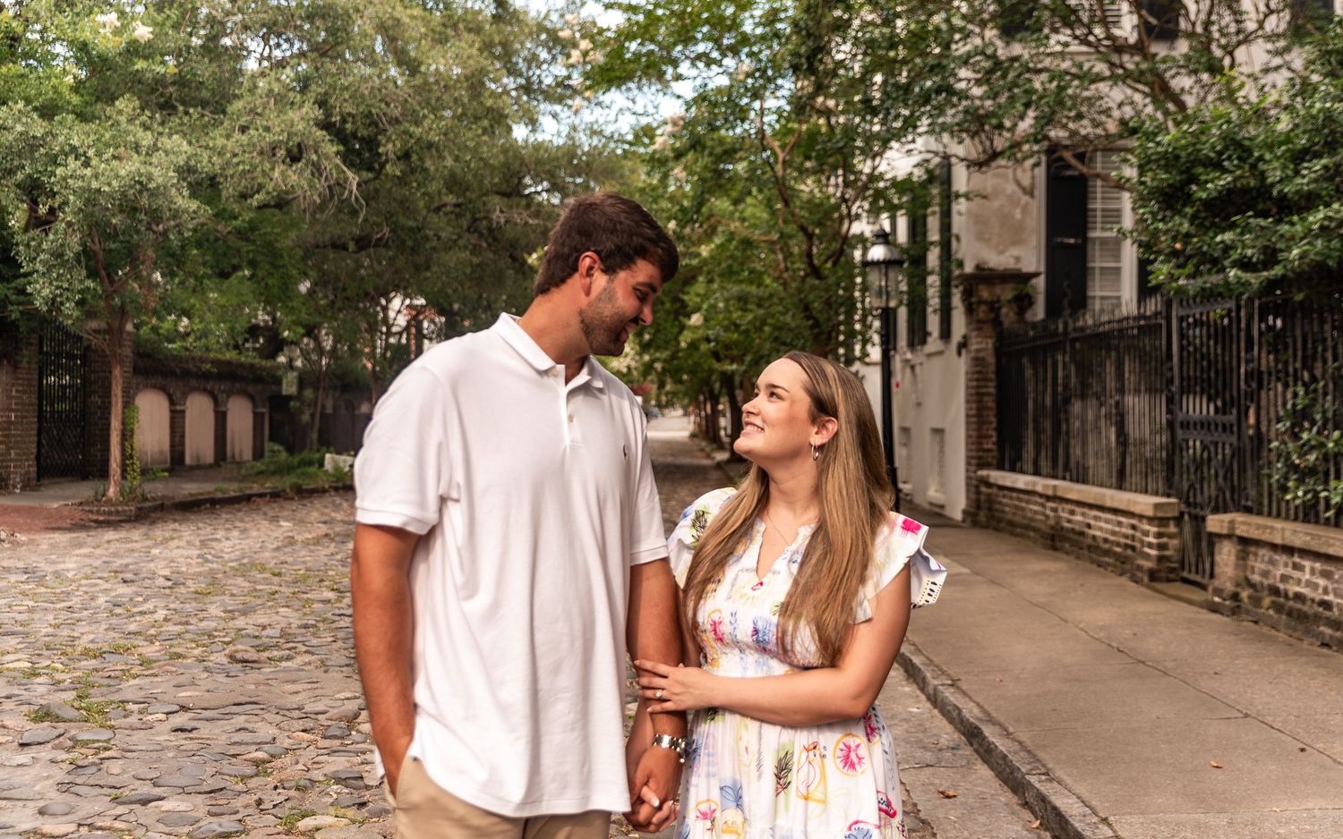 A couple shares a romantic moment while walking along a cobblestone street on a sunny afternoon.