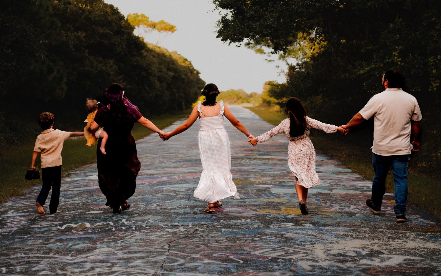 Group holds hands while walking along rocky beach at sunset creating silhouette effect.