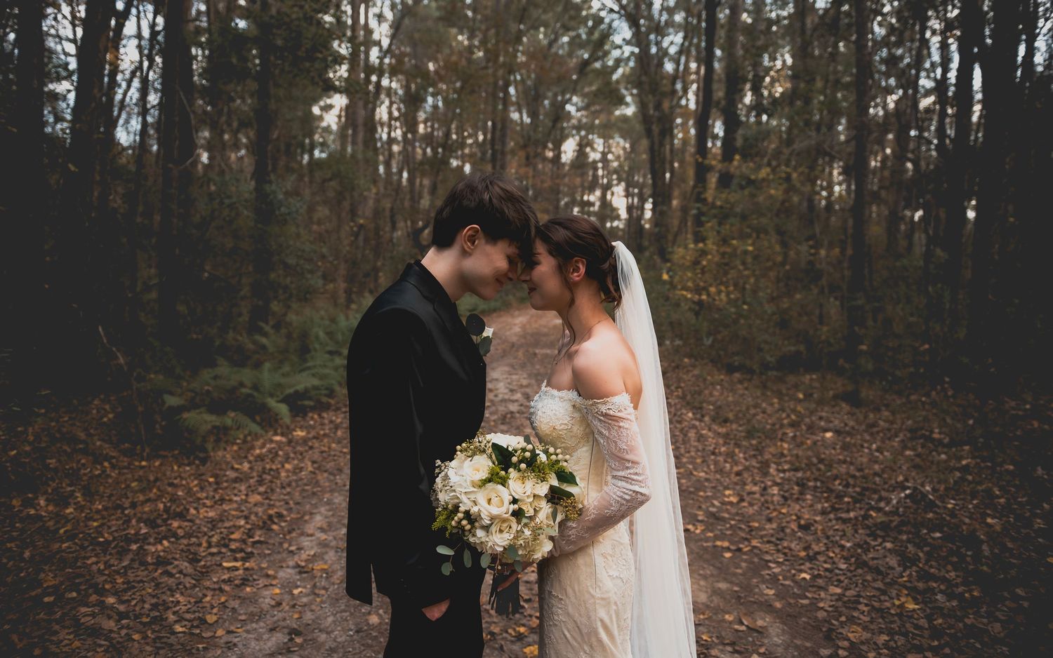 A romantic wedding couple shares an intimate moment in a moody autumn forest setting with fallen leaves.