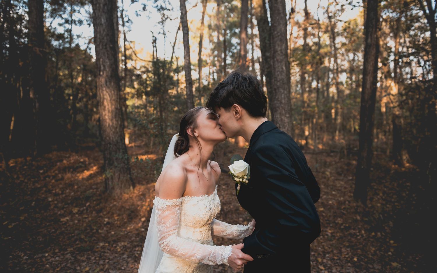 A romantic wedding couple shares an intimate moment in a sunlit autumn forest with fallen leaves on the ground.
