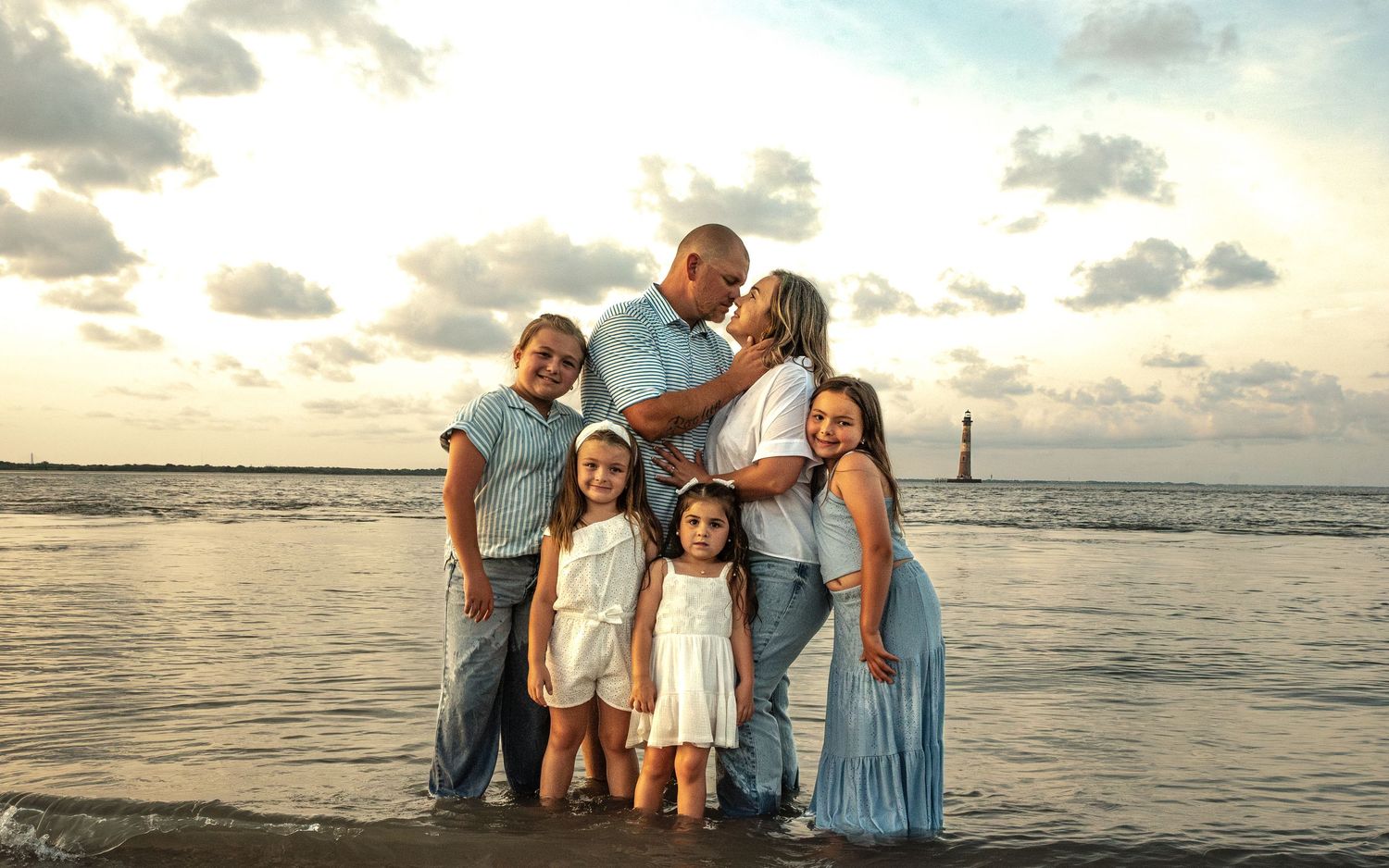 A family in coordinated blue and white outfits poses together on a scenic beach at sunset.