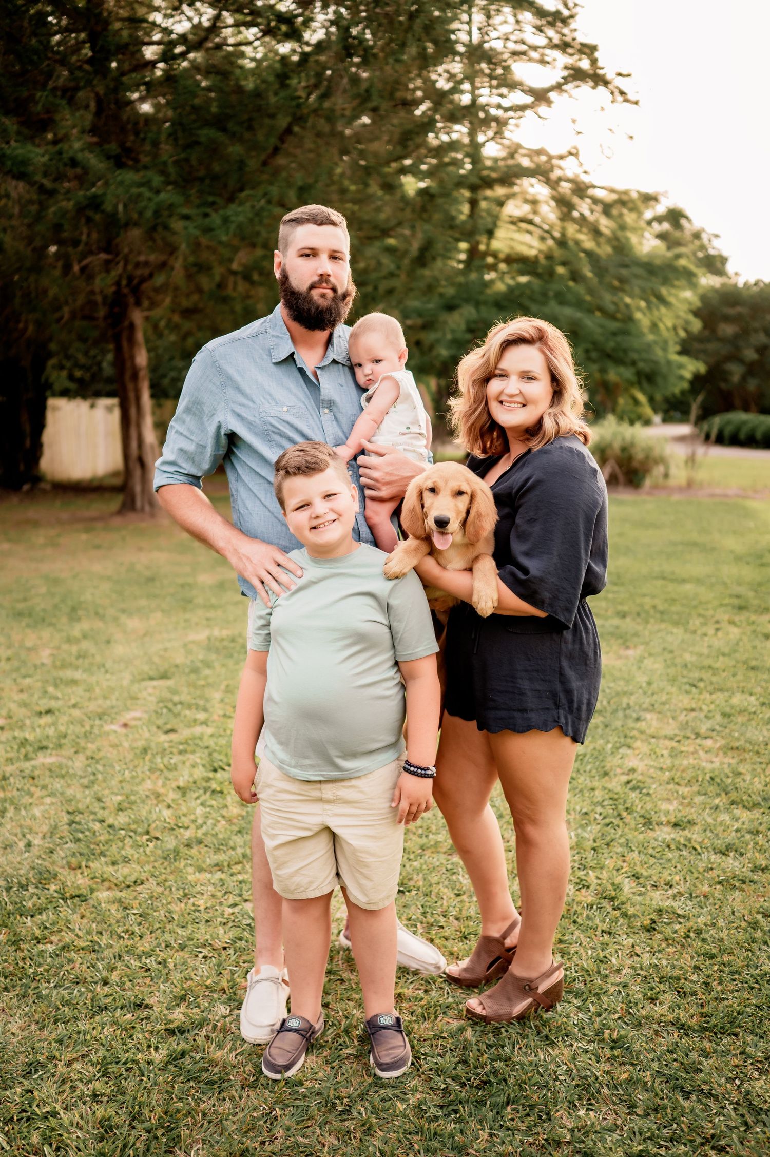 A family with their golden retriever dog poses together in a grassy outdoor setting at sunset.