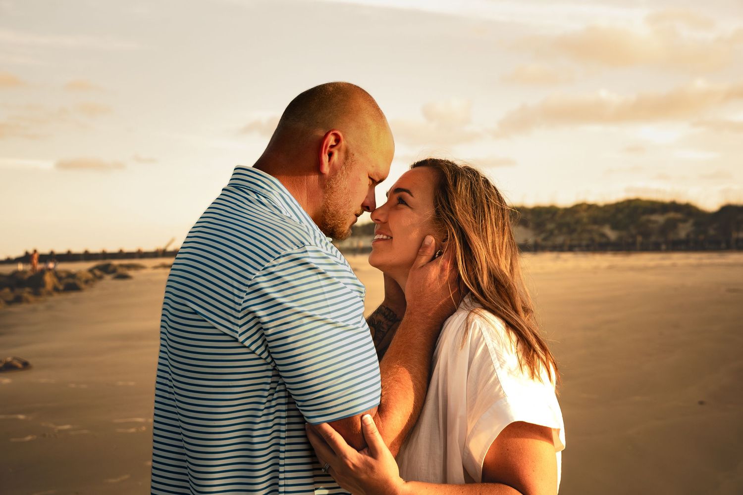 Couple in blue striped shirt and white top embracing on sandy beach at sunset.