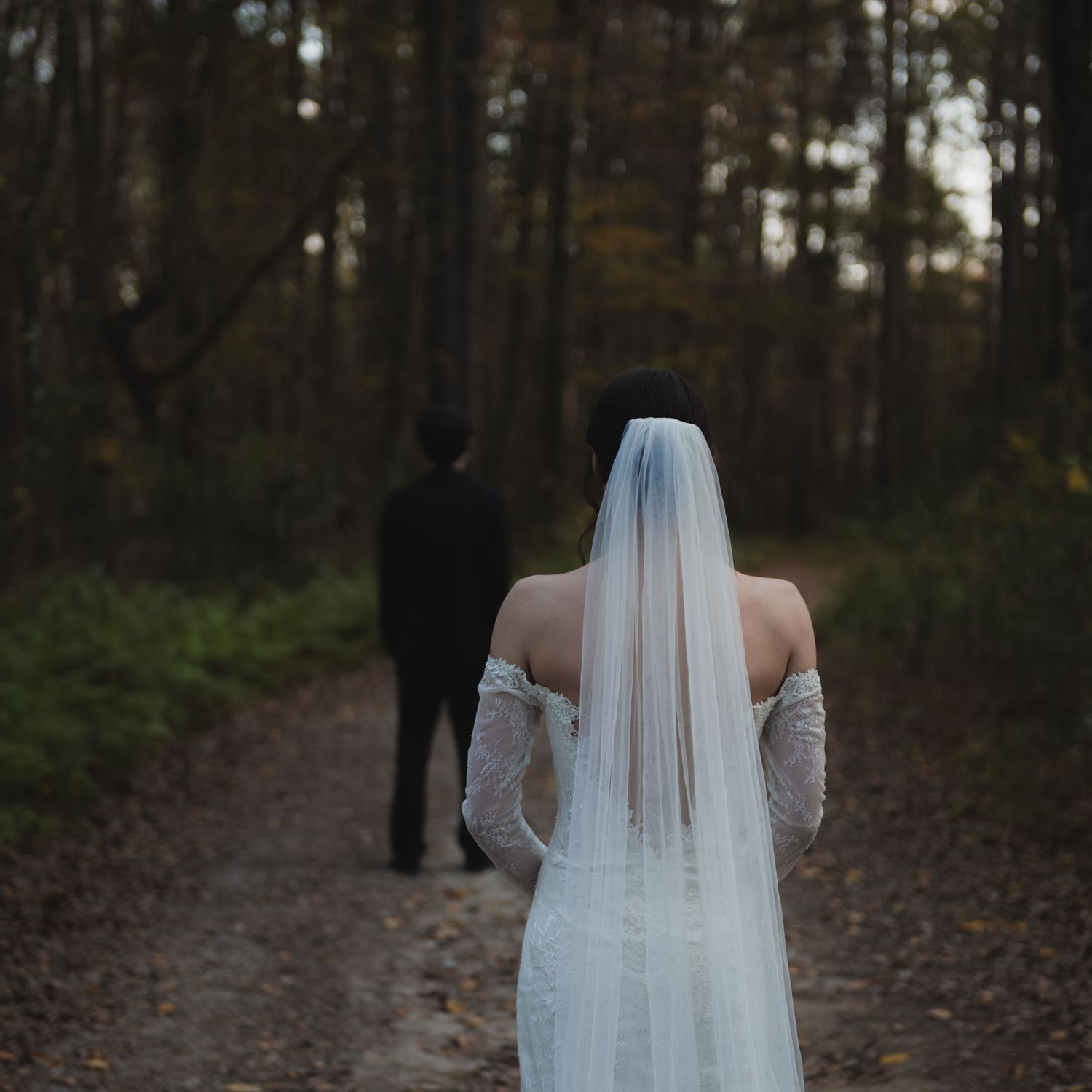 A bride in a flowing white wedding gown and veil walks through a wooded forest path during an autumn wedding.