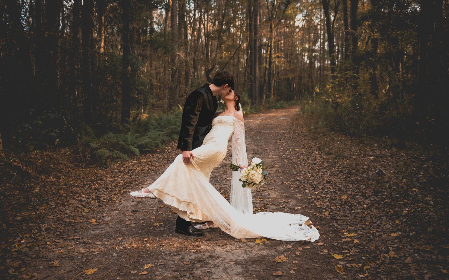 Romantic autumn wedding photos showing couple in white dress and suit posing on leaf-covered forest path at sunset.