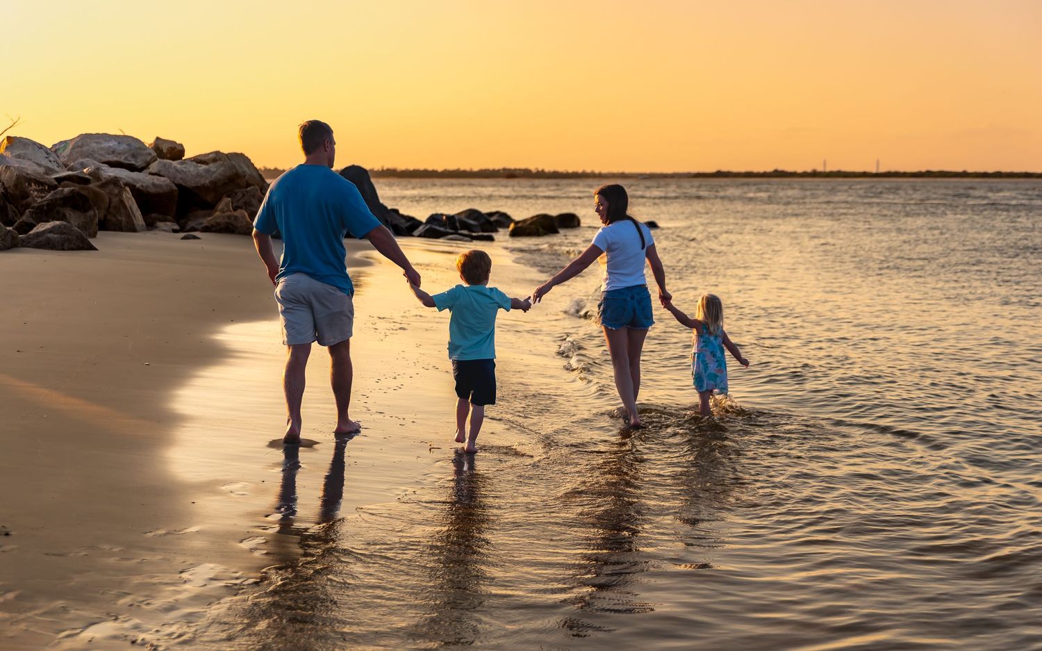 A family walks along the beach shoreline at sunset creating silhouettes against the golden sky and water.